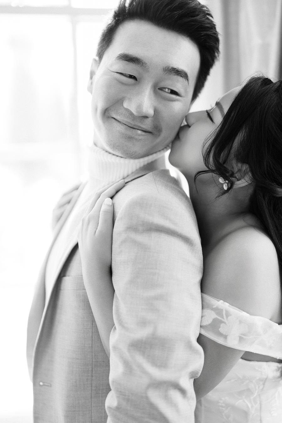 A romantic black and white photo of a couple embracing, with the woman gently kissing the man's cheek, capturing a tender moment of affection.