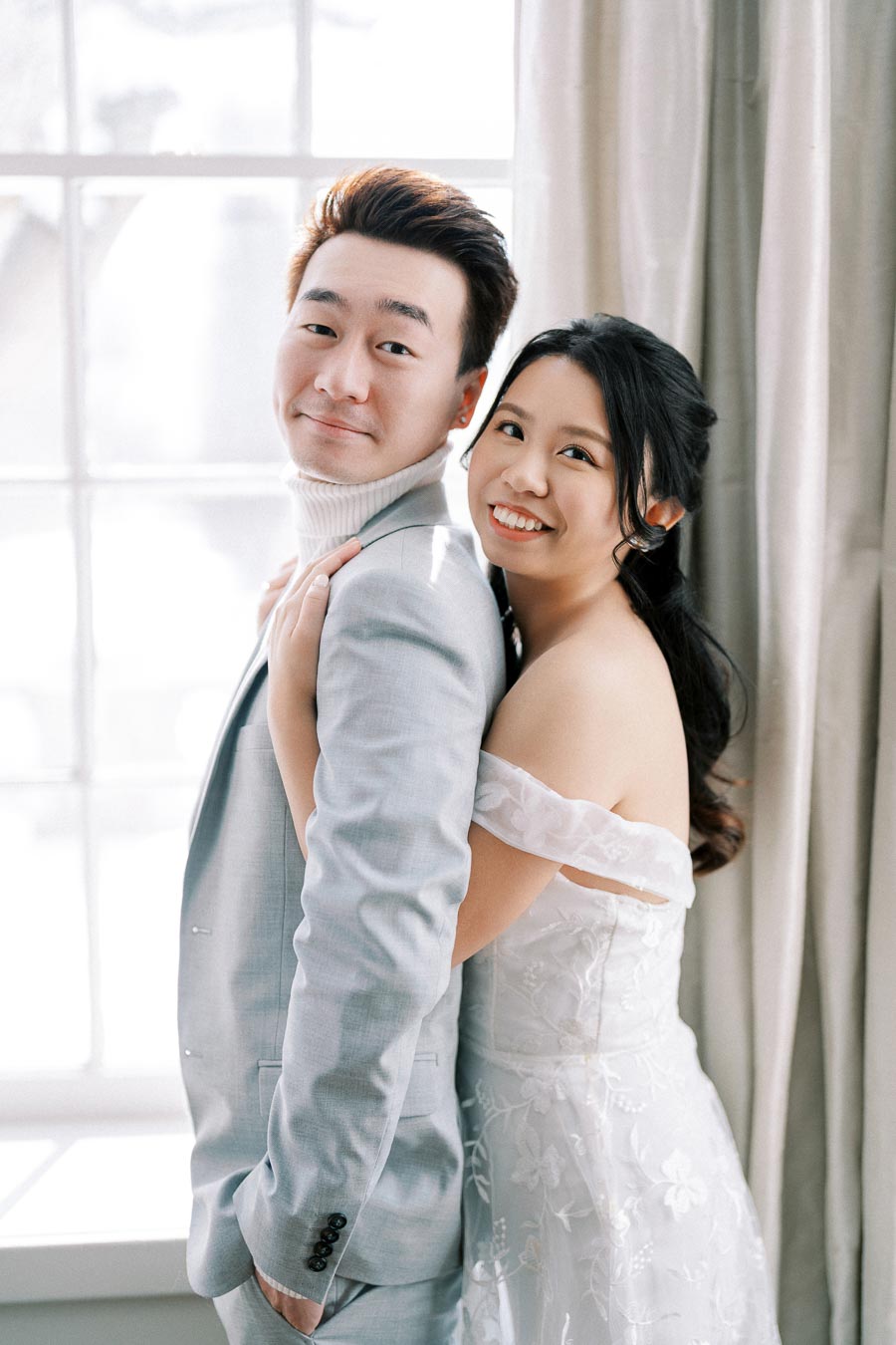 Smiling couple embracing in wedding attire near a bright window, showcasing love and happiness.