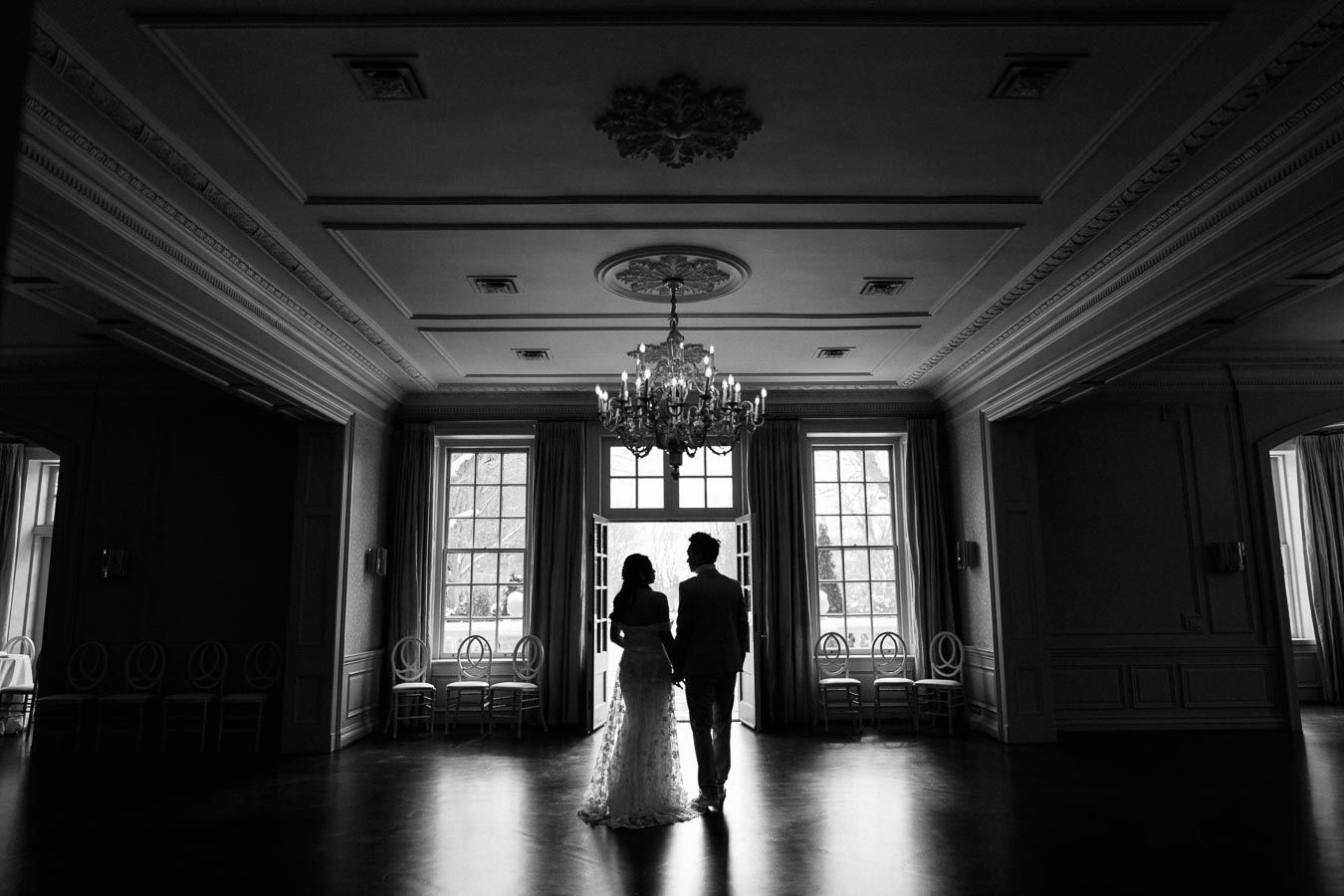 A couple stands in a grand, elegantly lit ballroom, silhouetted against a large window with an ornate chandelier above, creating a romantic and timeless ambiance.