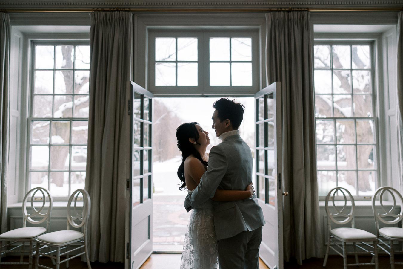 A romantic couple embracing near large windows in a sunlit room, with snow-covered trees visible outside.