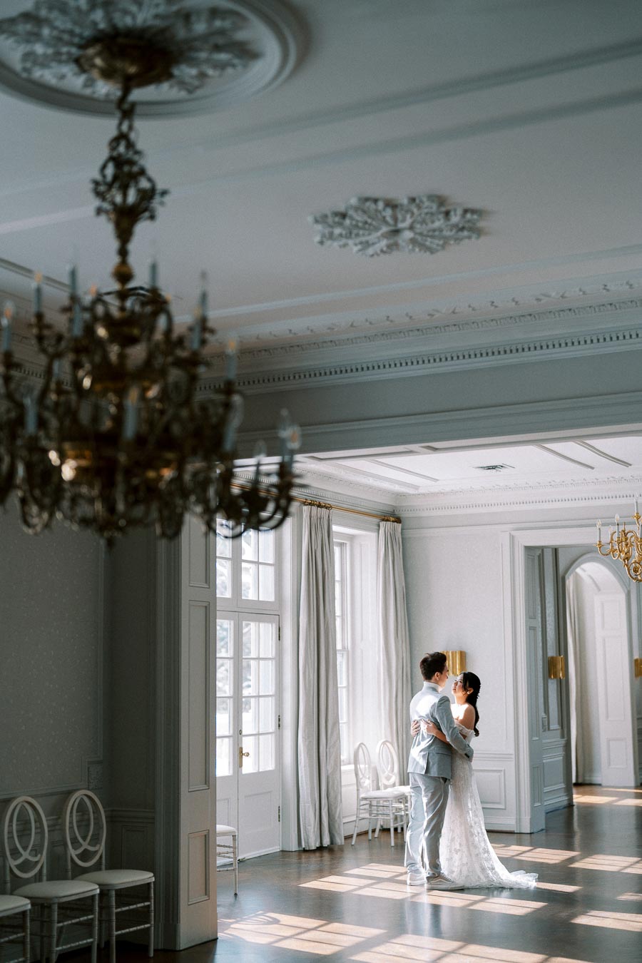 A couple dances in a sunlit, elegant ballroom with high ceilings, large windows, and ornate chandeliers, capturing a romantic wedding moment.