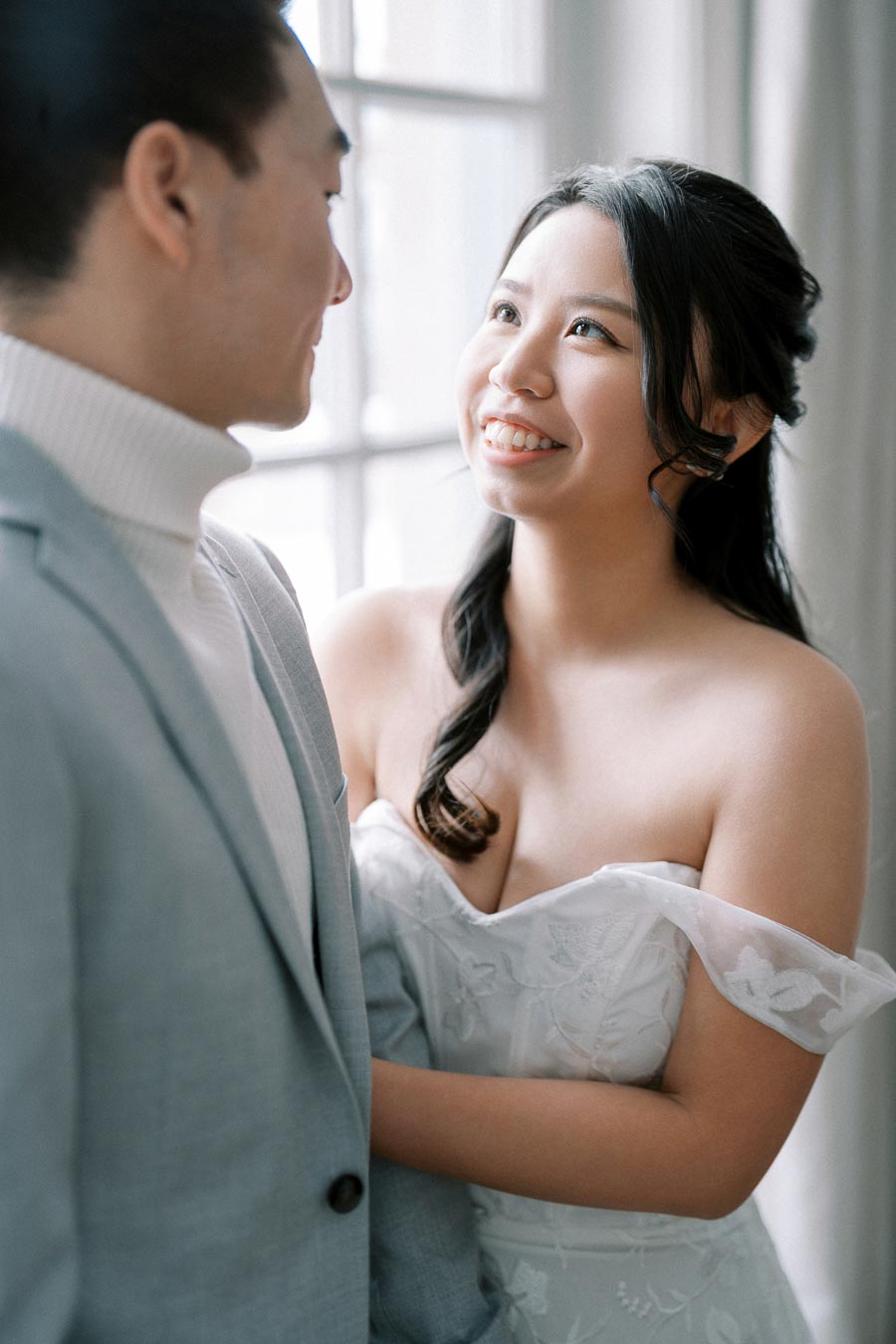 Happy couple gazing at each other in a bright room, with the woman in a white dress and the man in a light gray suit, capturing a romantic and tender moment.