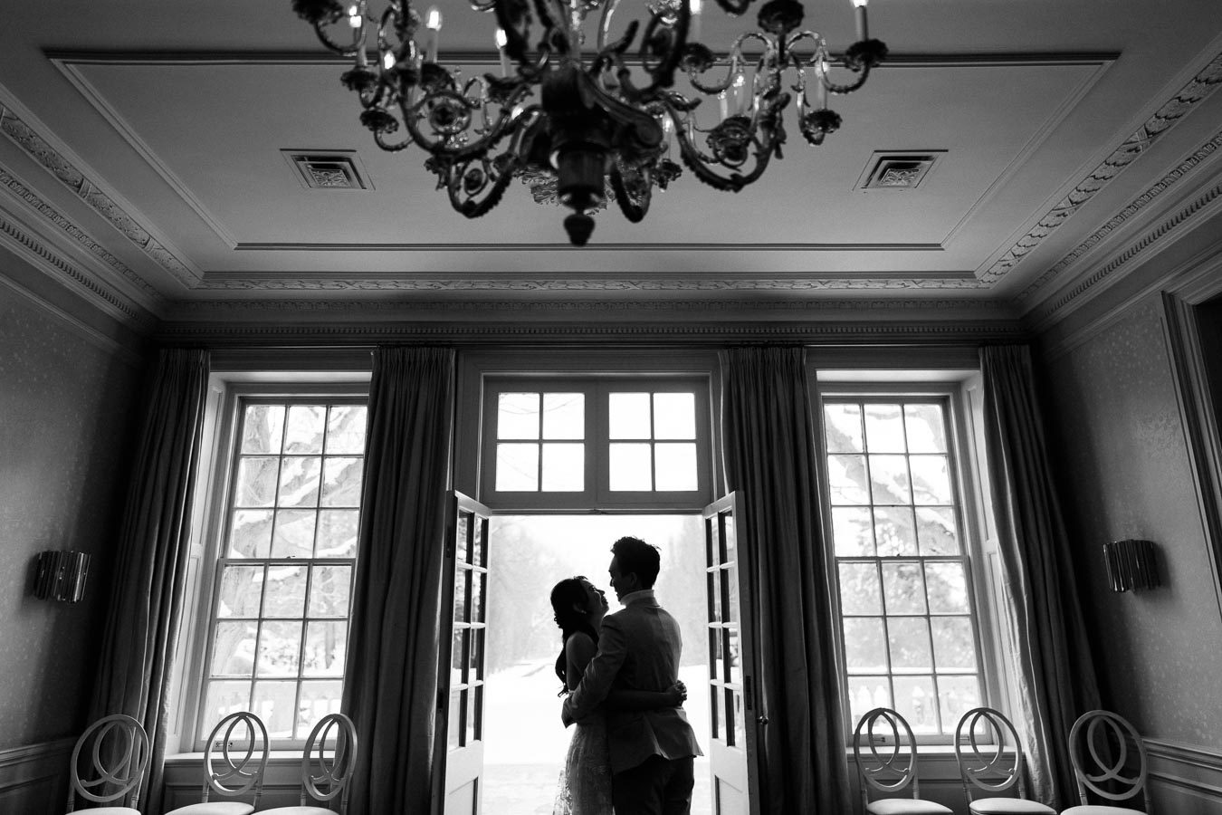 A bride and groom share a romantic embrace in a beautifully decorated room with large windows and an elegant chandelier, creating a timeless black and white wedding scene.