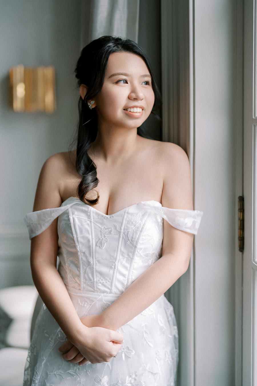 A bride in a white, strapless wedding gown with floral details, standing by a window and smiling, with natural light illuminating the scene.