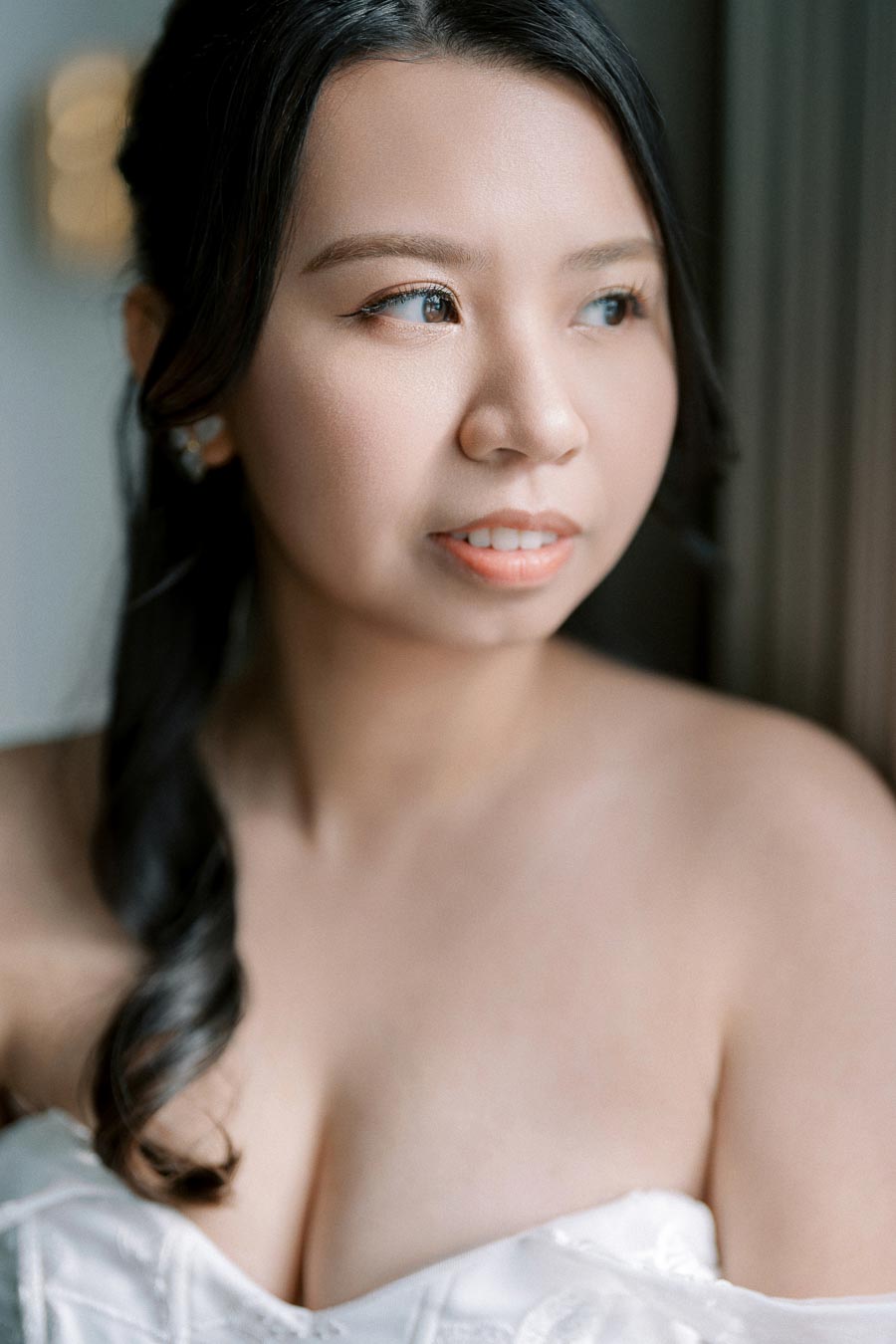 A woman in a white dress gazes thoughtfully out of a window, with soft lighting highlighting her features.