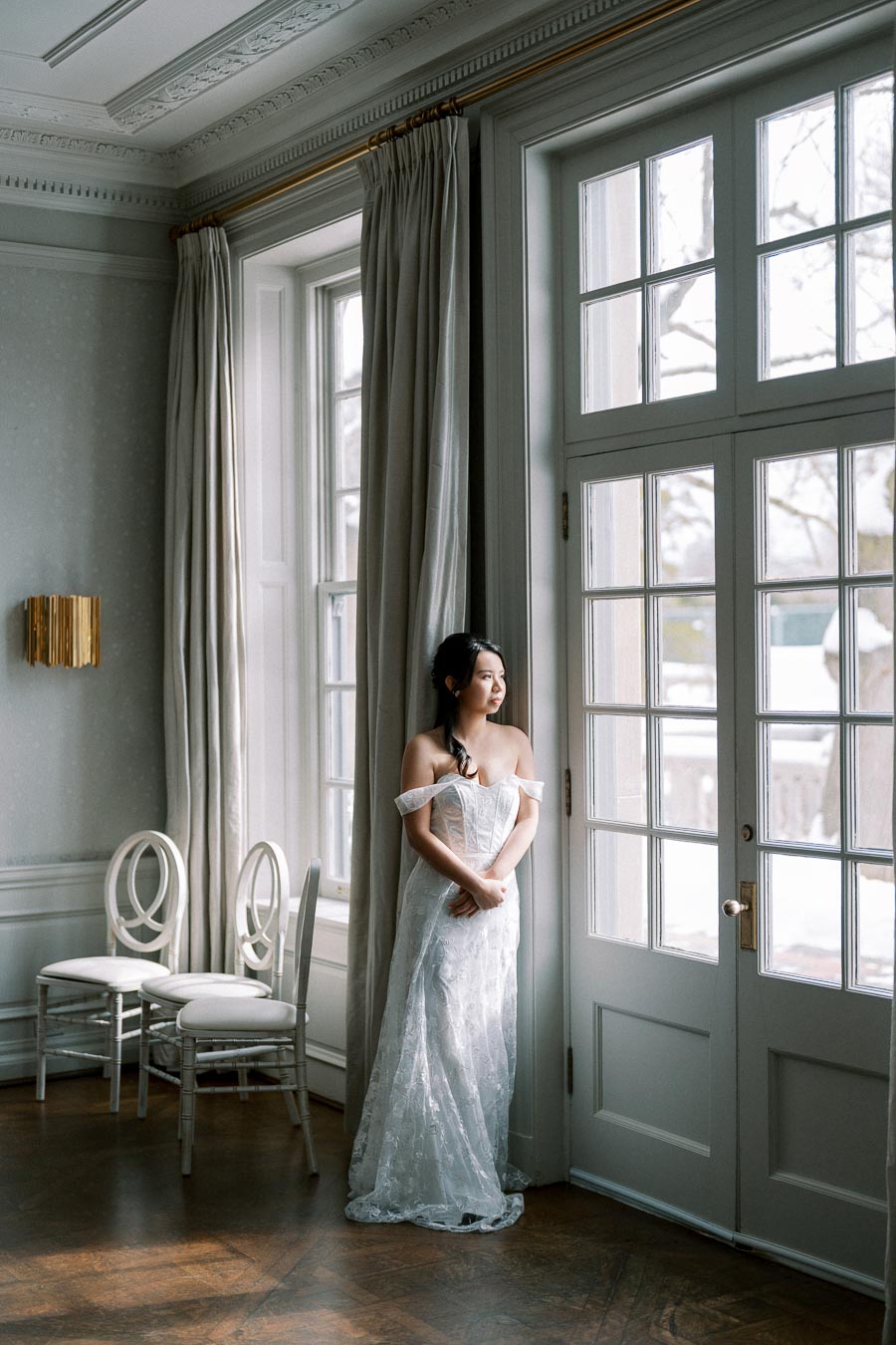 Bride in elegant white gown standing near a large window in a classic, white-walled room, looking outside thoughtfully.