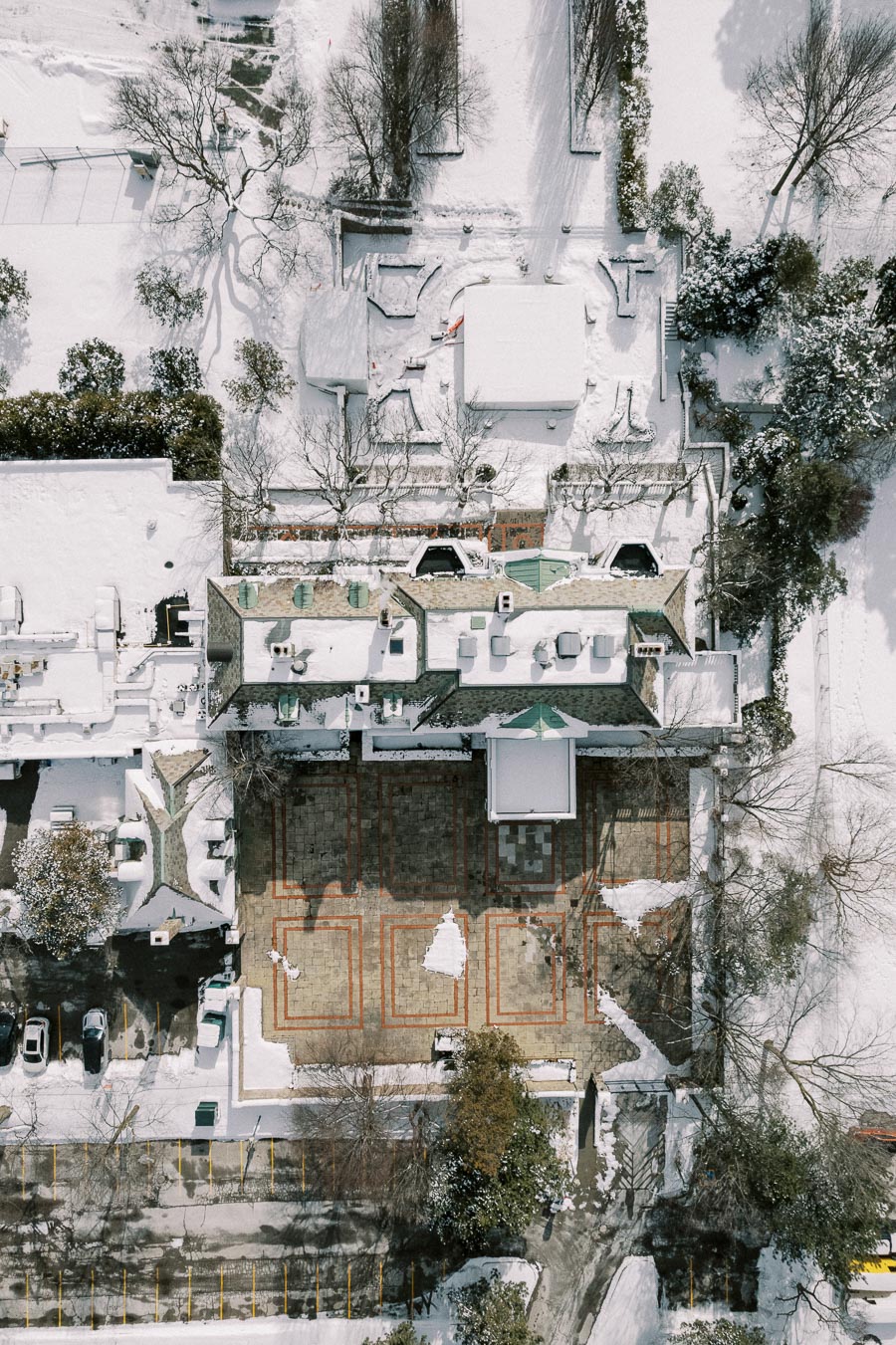 Aerial view of a snow-covered building complex with intricate roof patterns and surrounding trees, showcasing a large, partially cleared courtyard and nearby parking area.