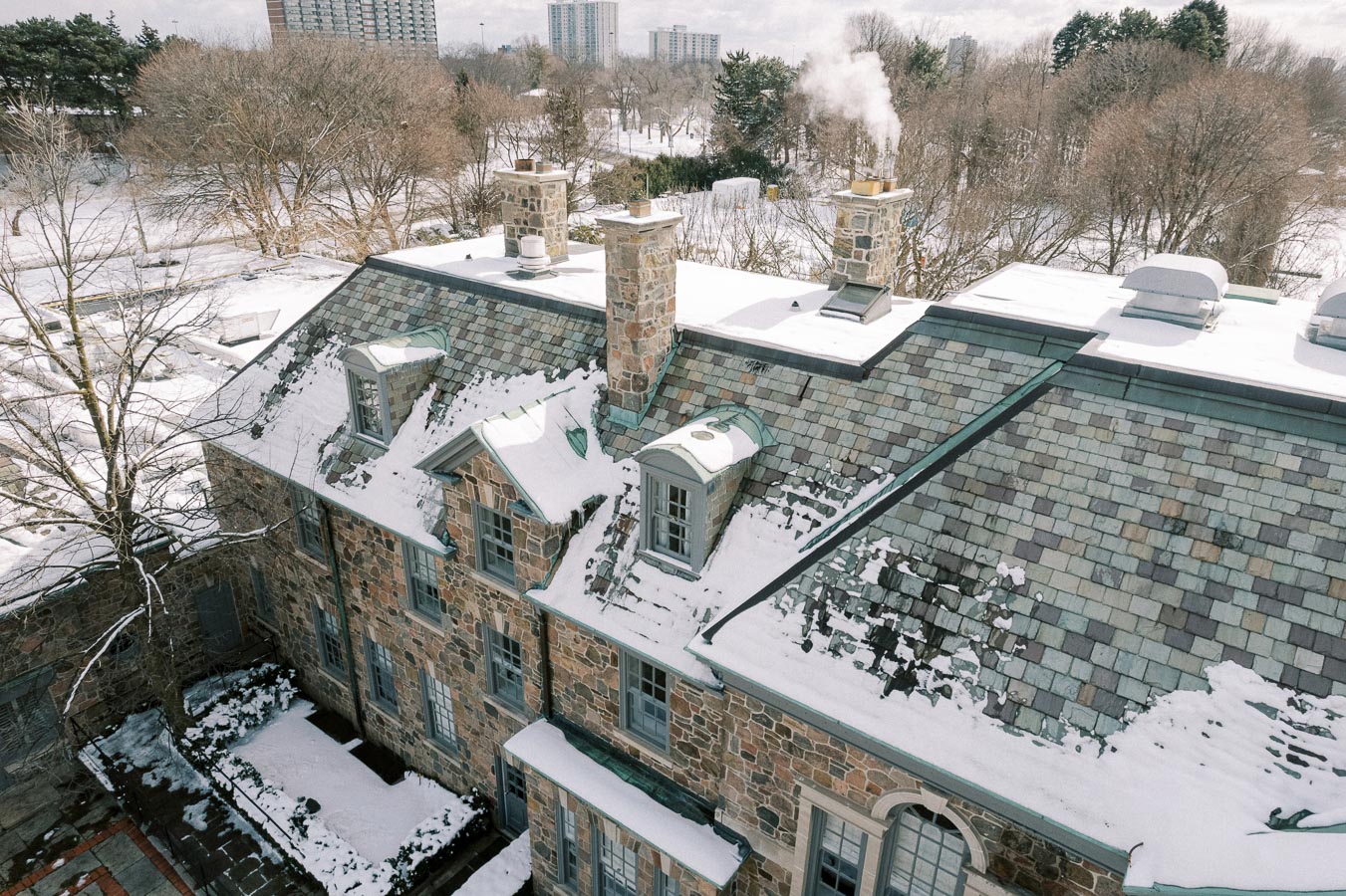 Aerial view of a historic stone building with a snow-covered roof and chimneys, surrounded by snowy trees and urban high-rise buildings in the background.