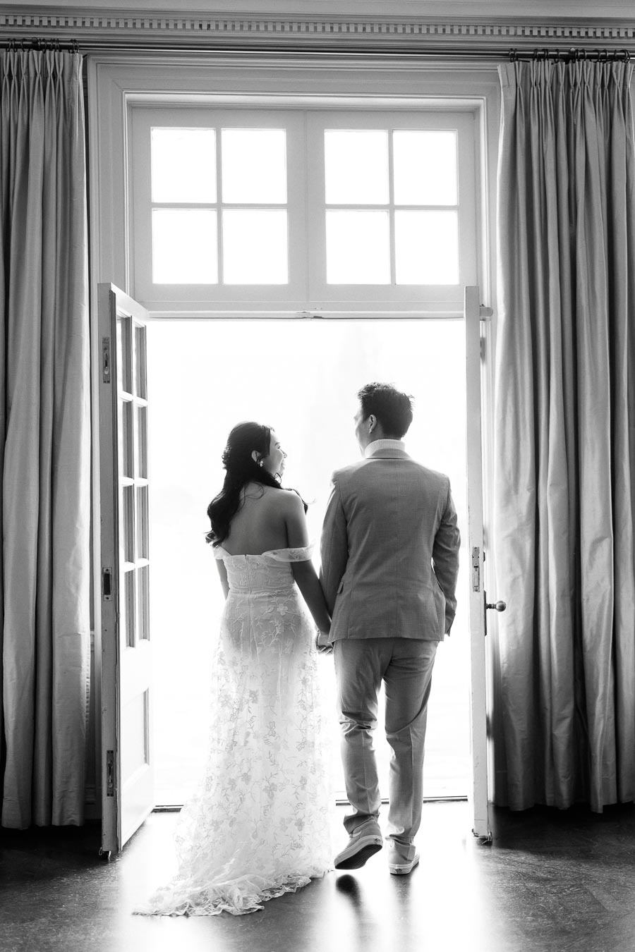 Black and white image of a couple holding hands and standing in a doorway, the bride in a lace wedding dress and the groom in a suit, gazing at each other.