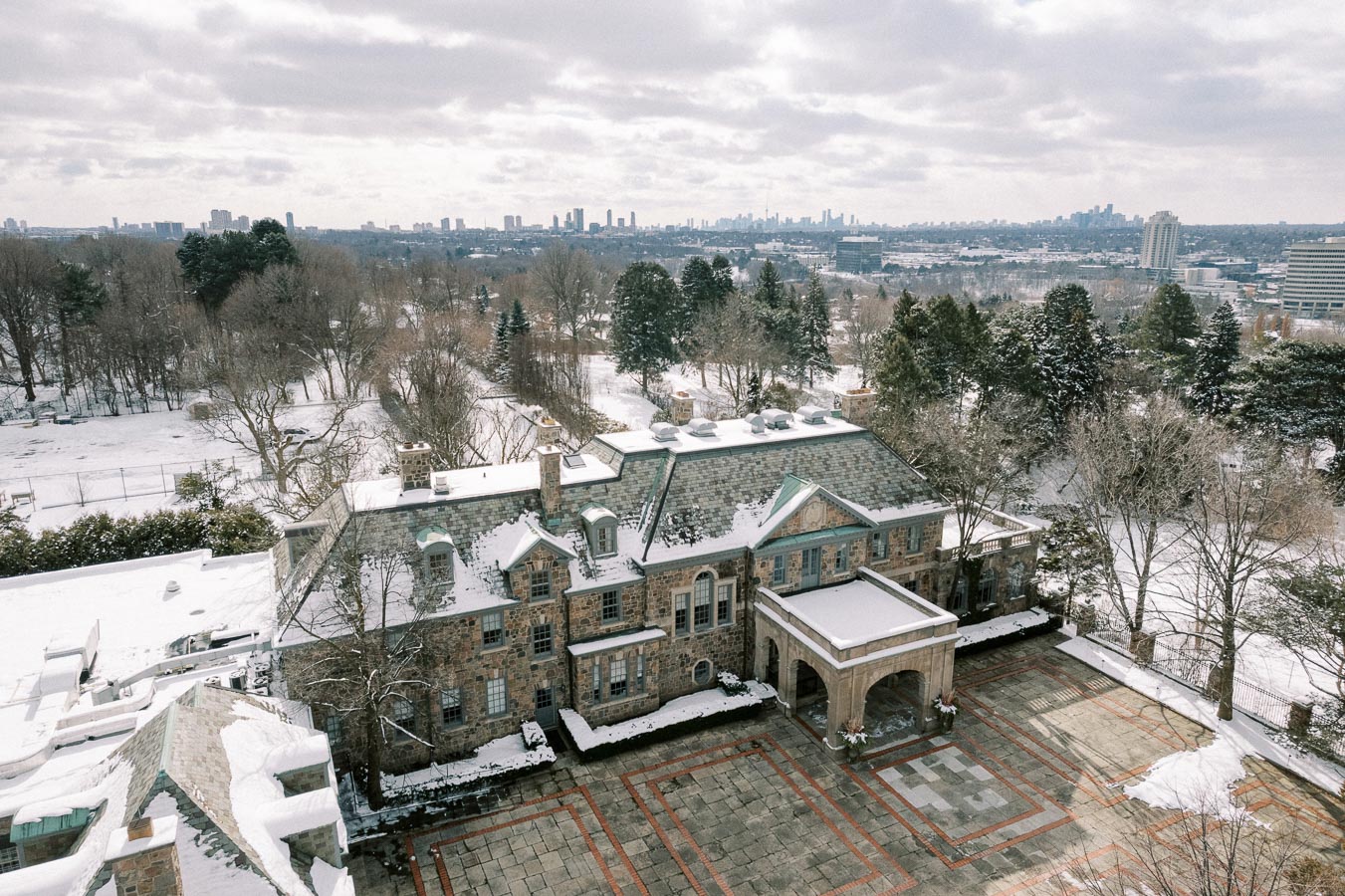 Aerial view of historic stone mansion covered in snow, surrounded by winter landscape and trees, with city skyline visible in the distance under a cloudy sky.