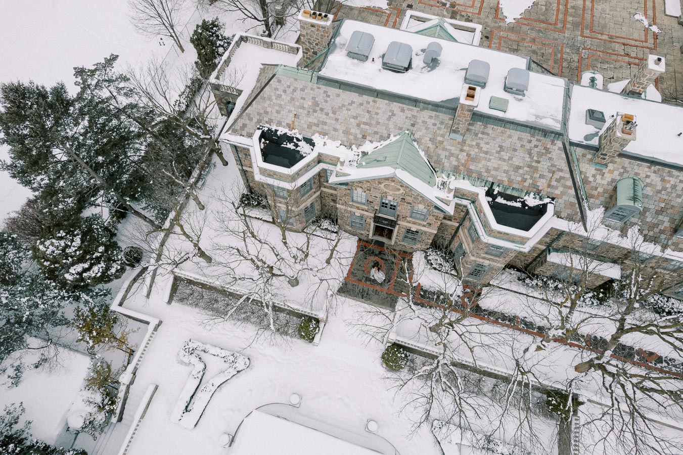 Aerial view of a snow-covered elegant stone mansion with intricate architectural details, surrounded by bare trees and a neatly maintained garden blanketed in snow.