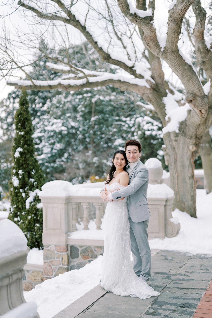 A couple dressed in wedding attire embraces in a snowy garden, with bare trees and a stone balustrade in the background, creating a romantic winter wedding scene.