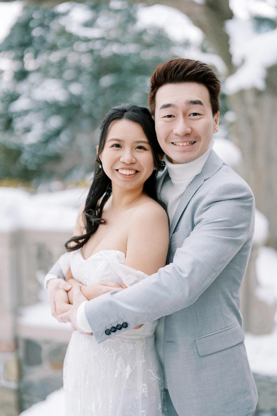 A happy couple embracing in a snowy outdoor setting, wearing formal attire. The woman is in a lace white dress, and the man is in a light gray suit with a white turtleneck.