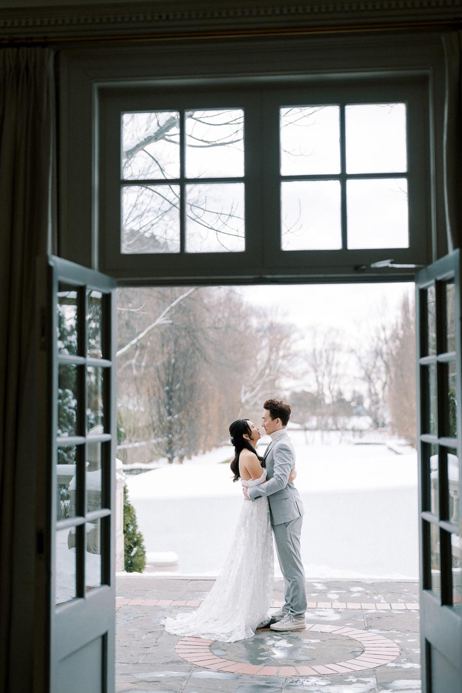 A couple embracing in a romantic winter wedding scene, viewed through open double doors with a snowy landscape in the background.