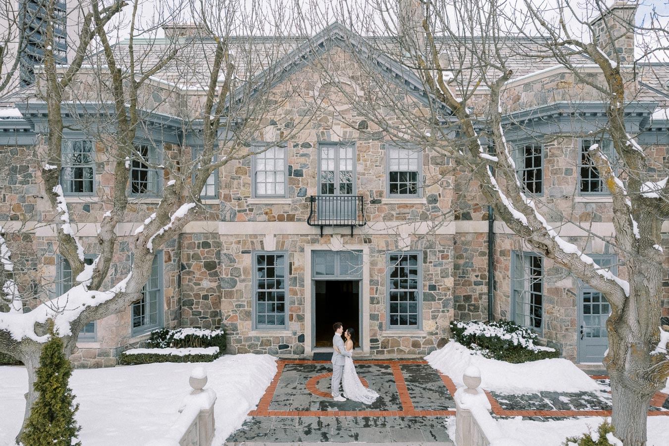Winter wedding couple standing outside a stone manor with snow-covered trees and pathway.