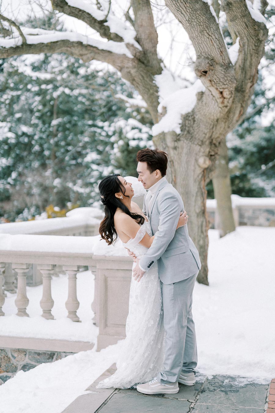 A couple in formal attire embracing in a snowy outdoor setting, with a stone railing and snow-covered trees in the background.