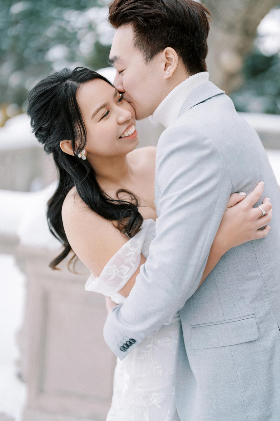 A happy couple embracing in a snowy outdoor setting, with the man kissing the woman's cheek, both dressed in elegant attire suitable for winter.