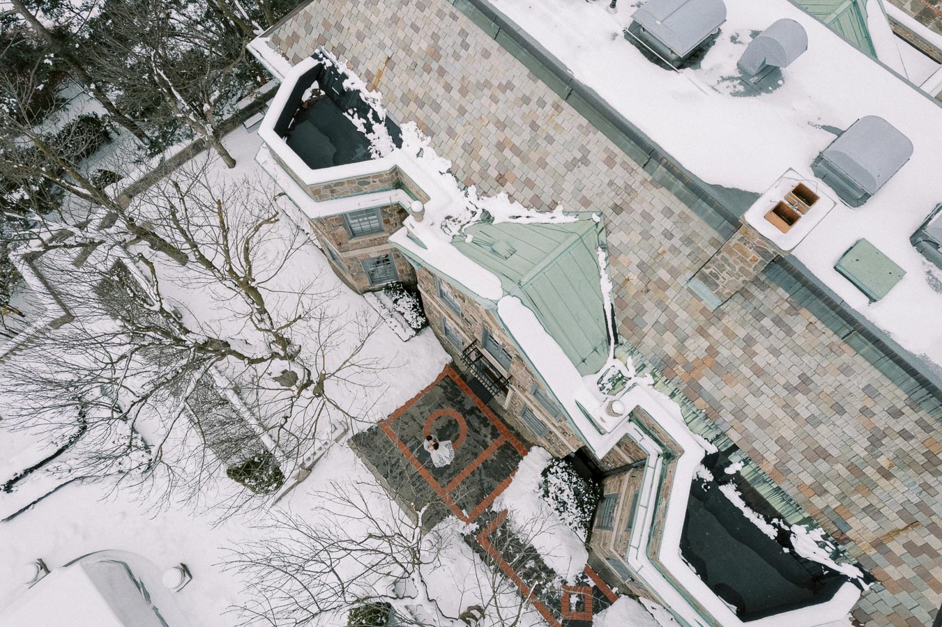 Aerial view of a snow-covered house with a patterned roof and courtyard surrounded by bare trees during winter.