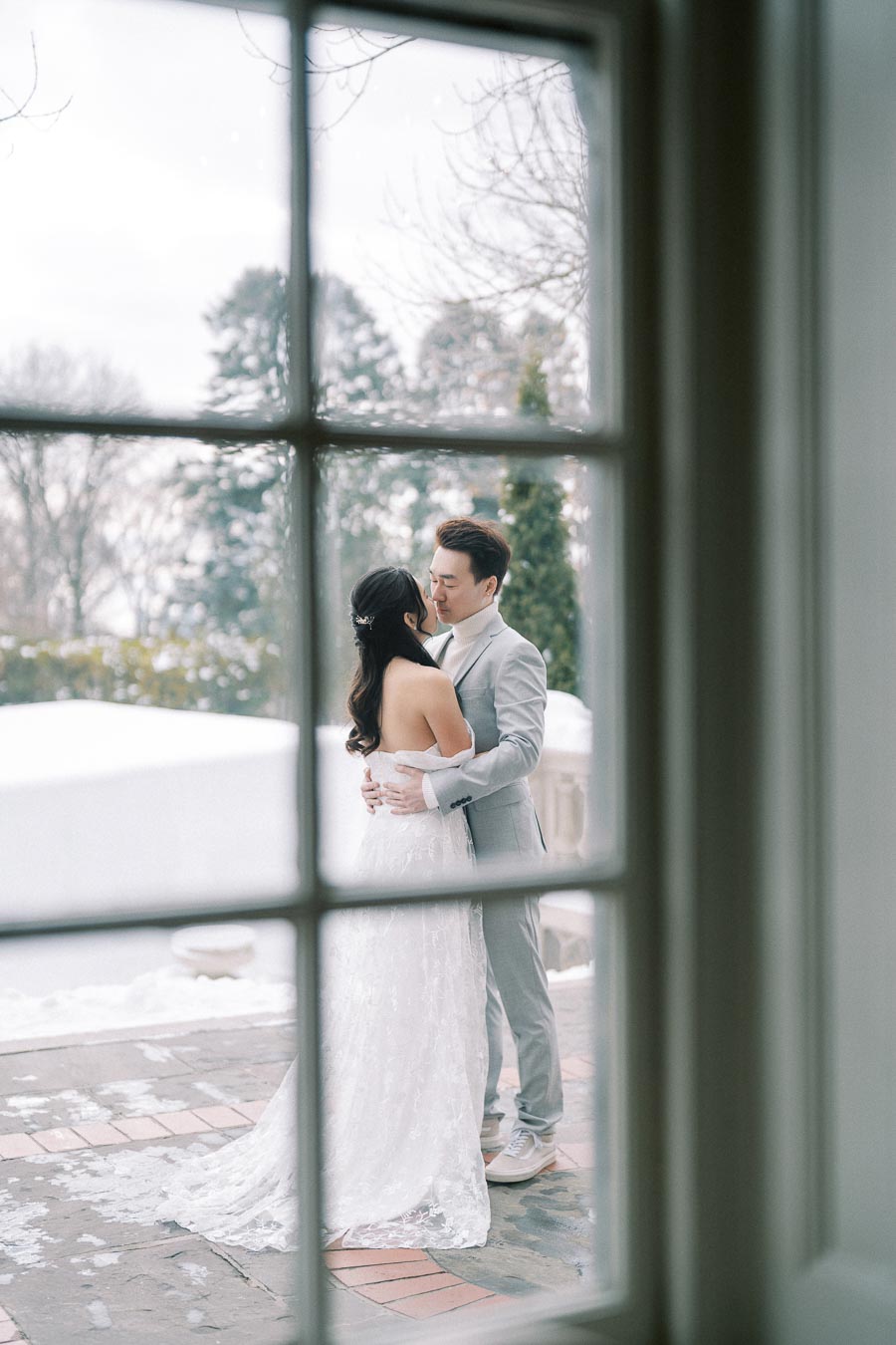 A bride and groom embracing on a snowy patio, viewed through a window, with winter trees in the background.