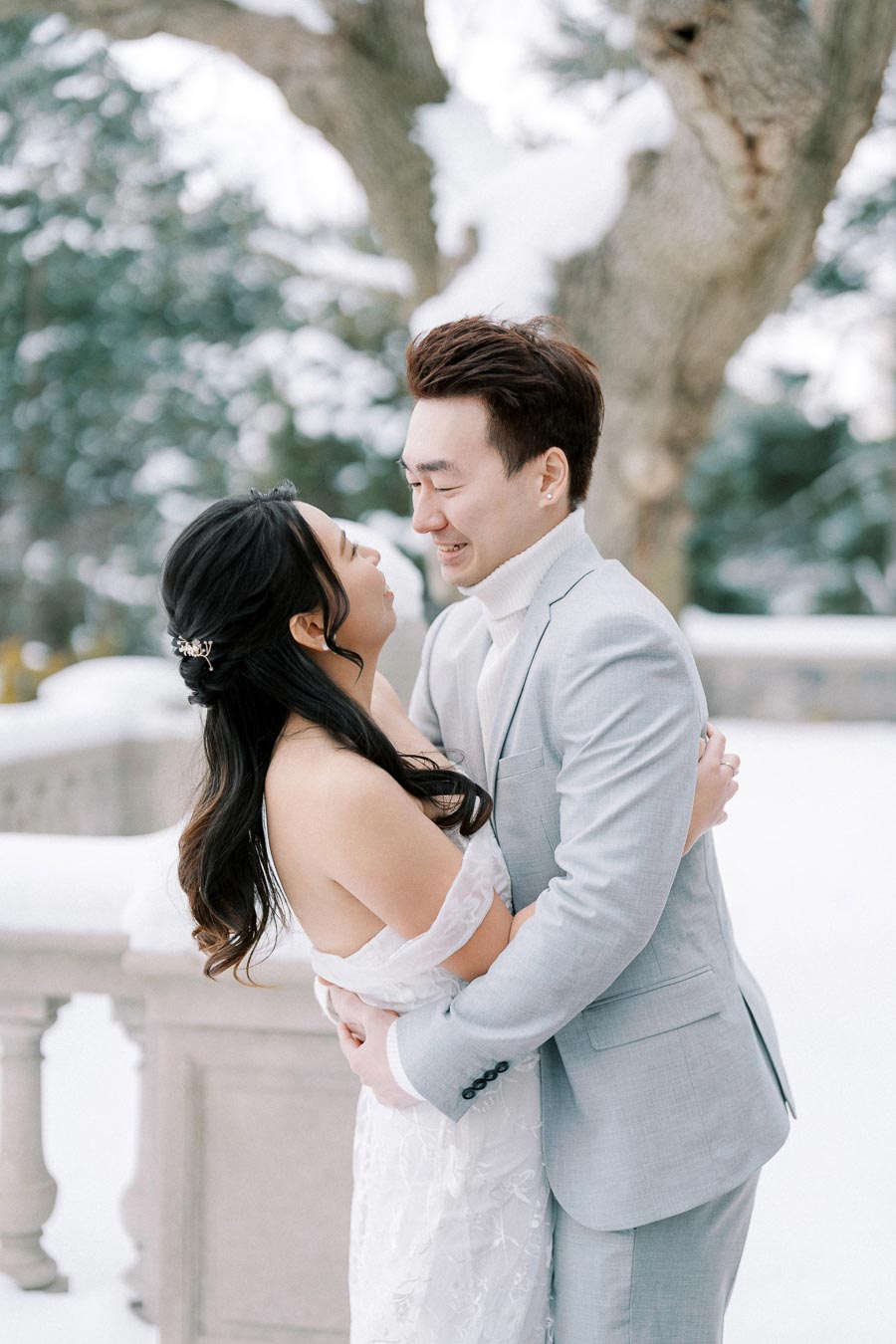 Romantic couple embracing in a snowy winter landscape, wearing elegant wedding attire, with a backdrop of snowy trees and a stone balustrade.