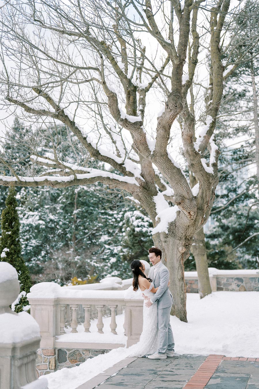 A couple in wedding attire embracing outdoors on a snowy winter day, standing near a large tree and stone railing.