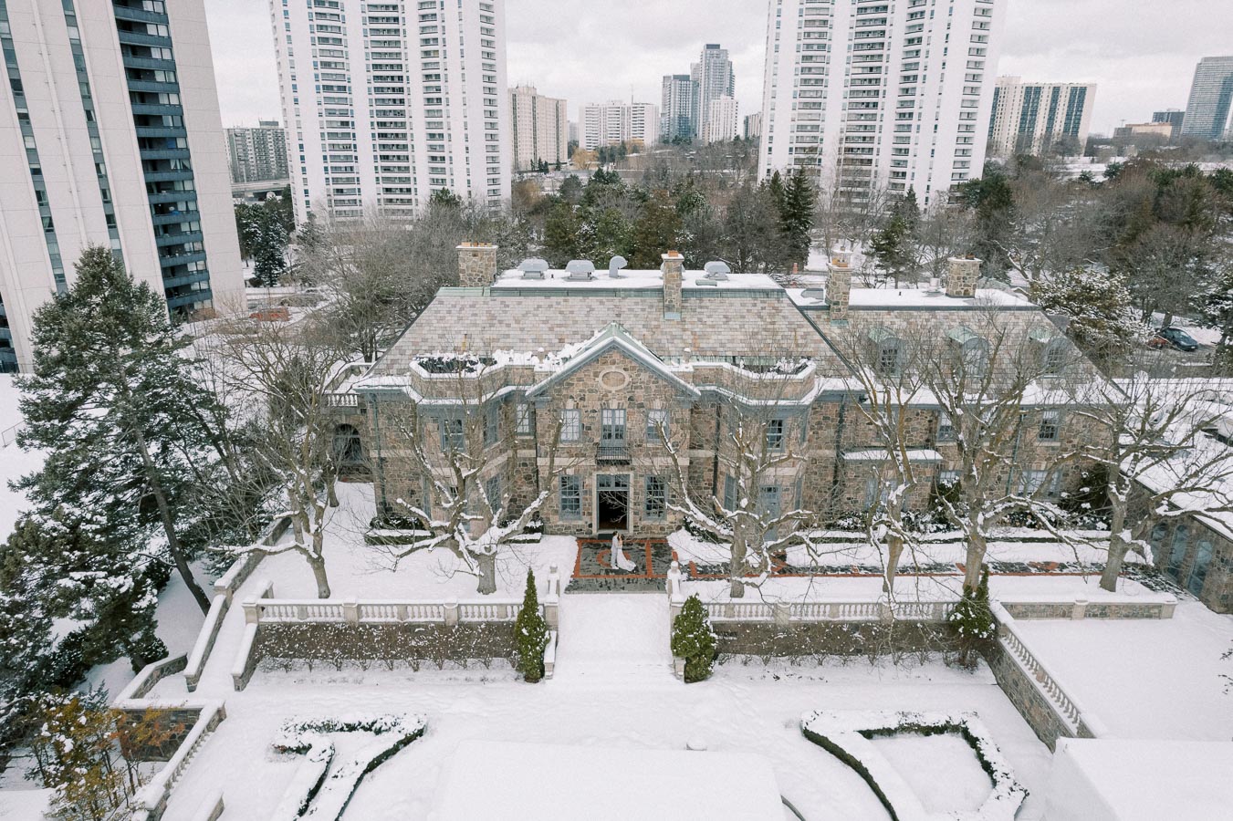 Snow-covered mansion surrounded by tall buildings in an urban winter setting, featuring bare trees and a classic architectural design.