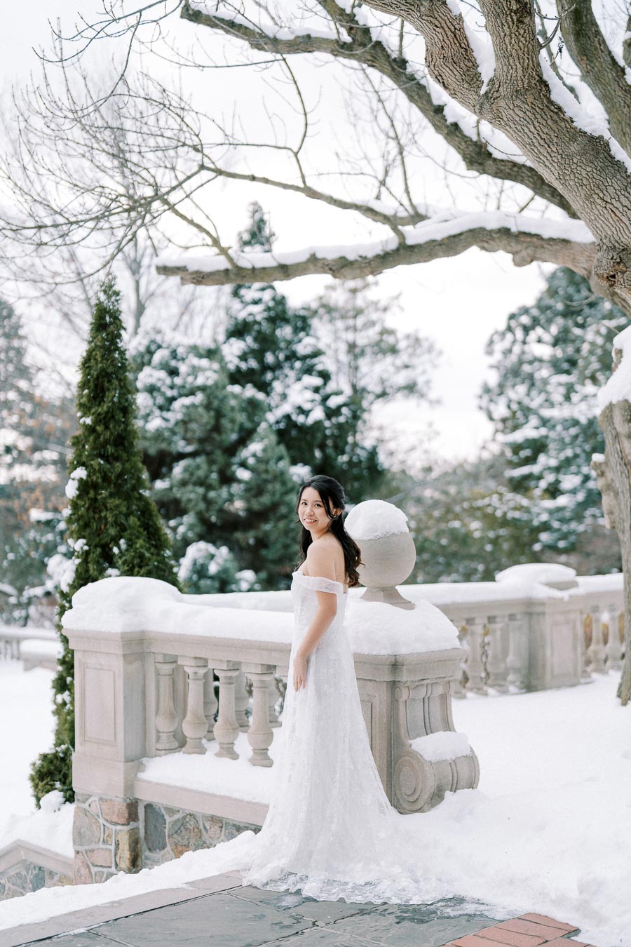 Bride in a white wedding dress standing next to a snow-covered stone balustrade, with winter trees and evergreens in the background.