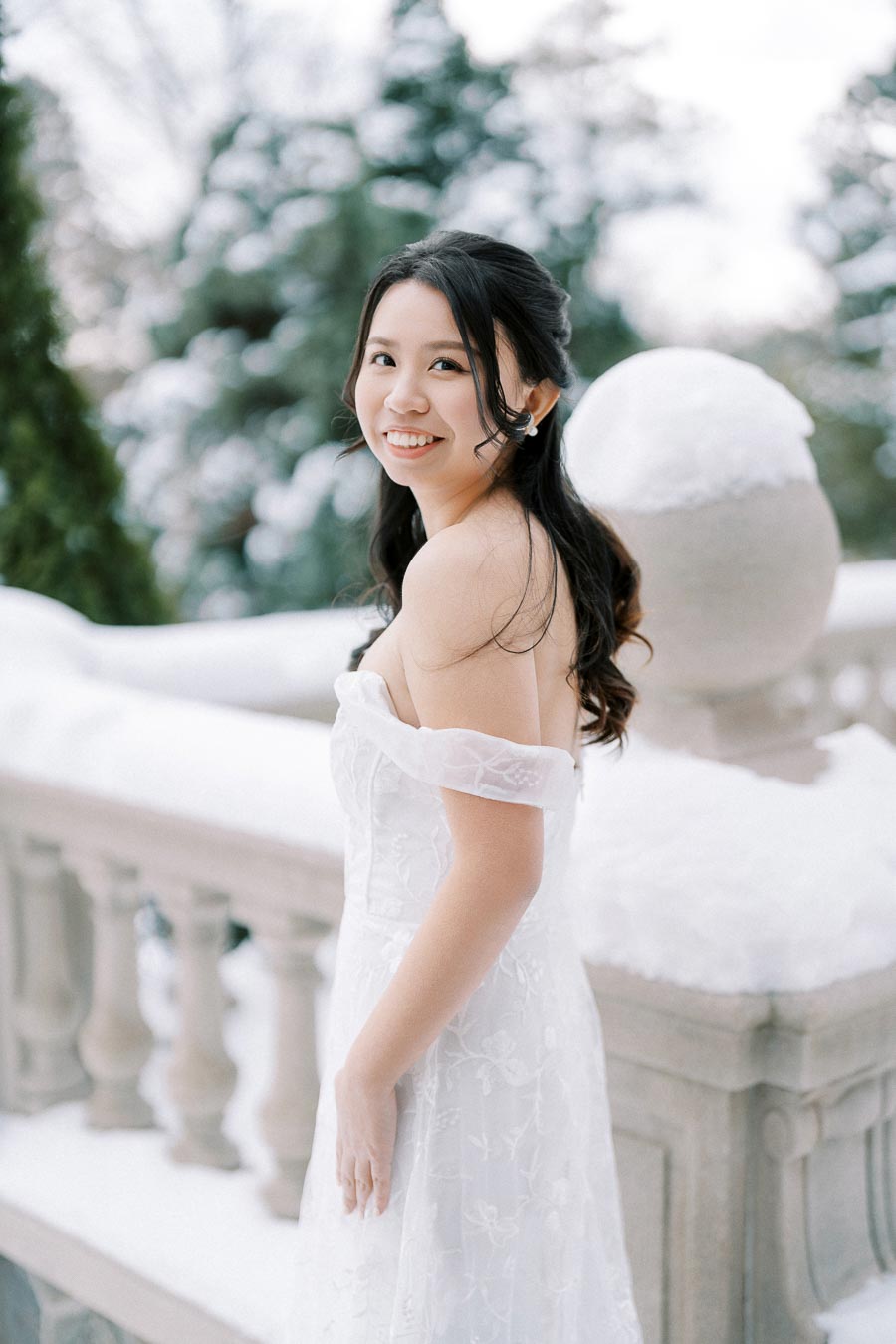 A woman in an elegant off-the-shoulder white dress smiles while standing on a snow-covered balcony with blurred snowy trees in the background.