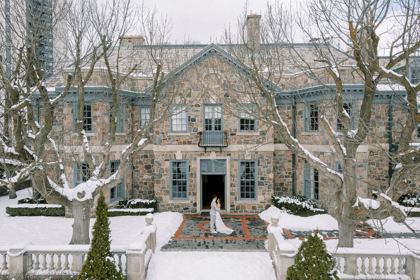Charming winter wedding scene in front of a historic stone building with snow-covered trees, featuring a couple embracing in elegant attire.