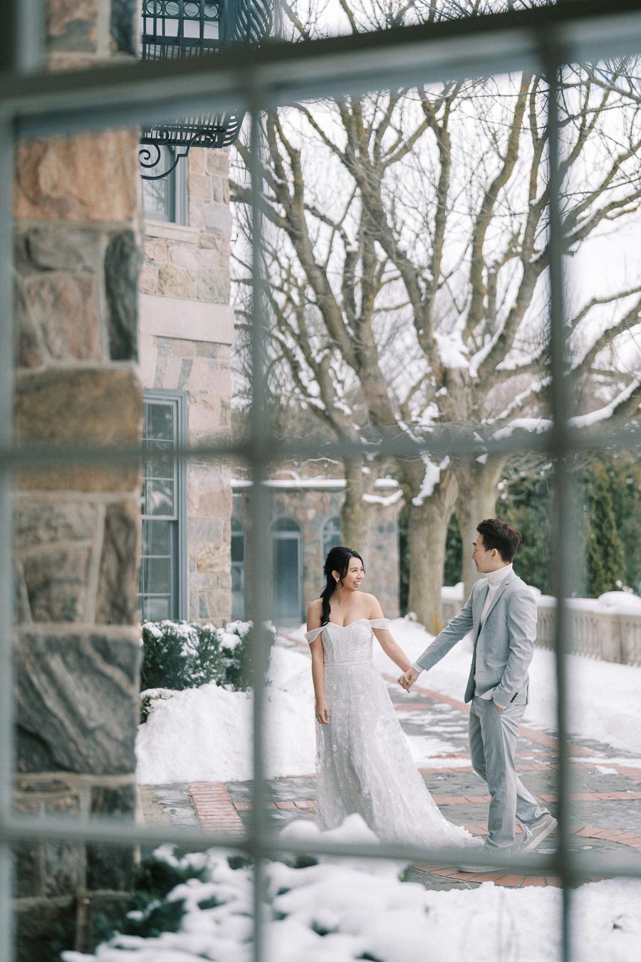 Winter wedding couple holding hands outside a stone building, surrounded by snow-covered trees and walkways, viewed through a window frame.