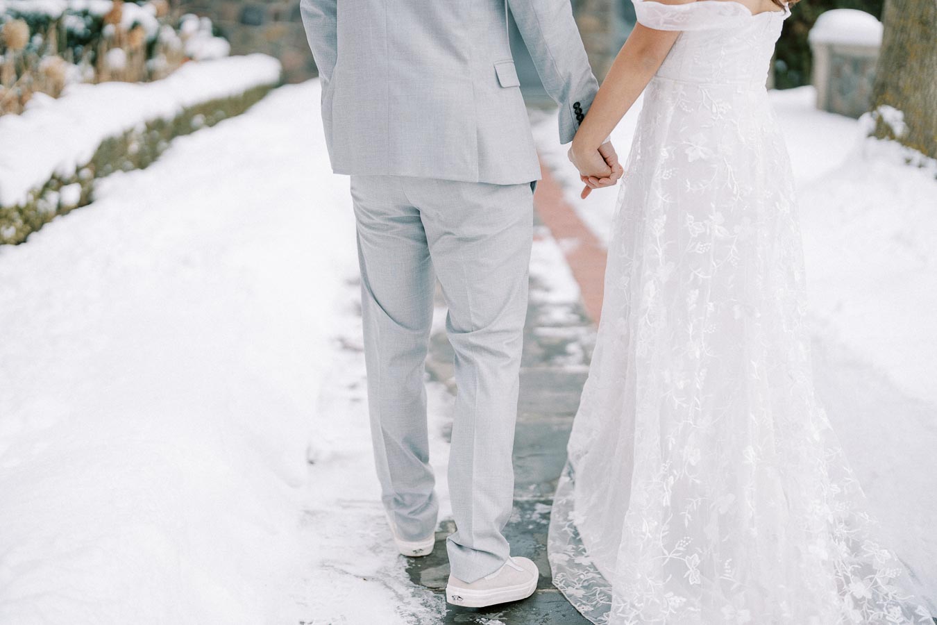 A bride and groom holding hands and walking down a snowy path; bride in a white lace dress and groom in a light gray suit with sneakers, symbolizing a winter wedding scene.