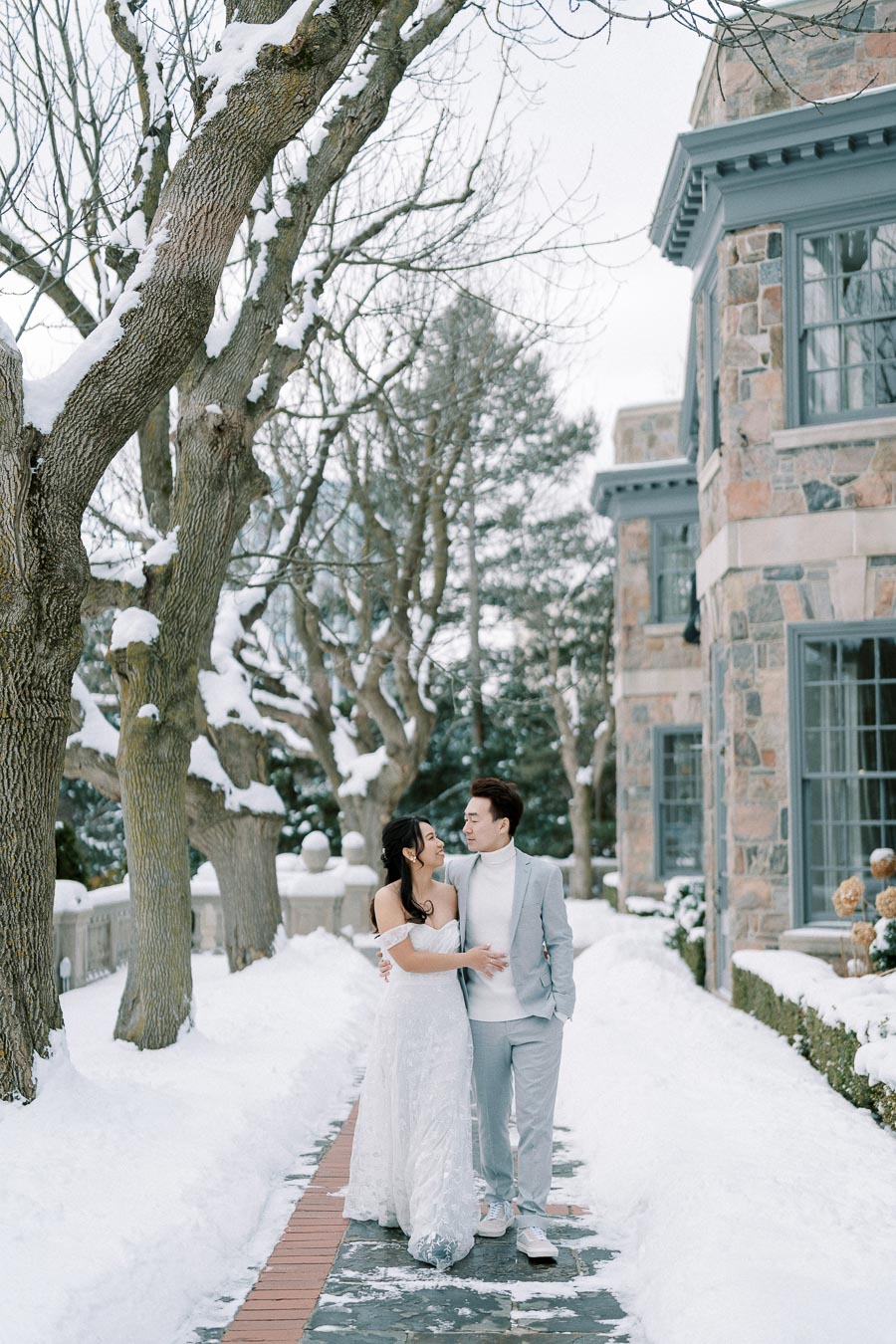 A couple in elegant winter attire stands joyfully on a snow-covered path lined with trees, in front of a charming stone building. The woman in a white dress and the man in a light grey suit share a loving gaze, surrounded by a serene winter landscape.