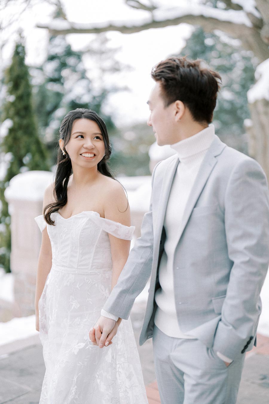 A bride in a white wedding dress and a groom in a gray suit holding hands and smiling at each other in a snowy outdoor setting.