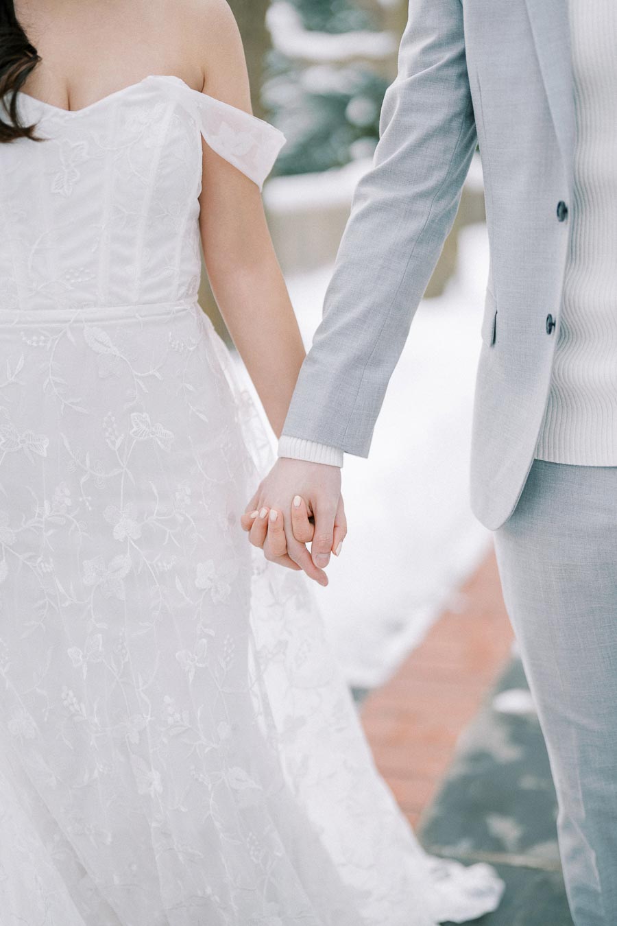A bride in an elegant lace wedding dress and a groom in a light gray suit holding hands during a winter wedding, with snow in the background.