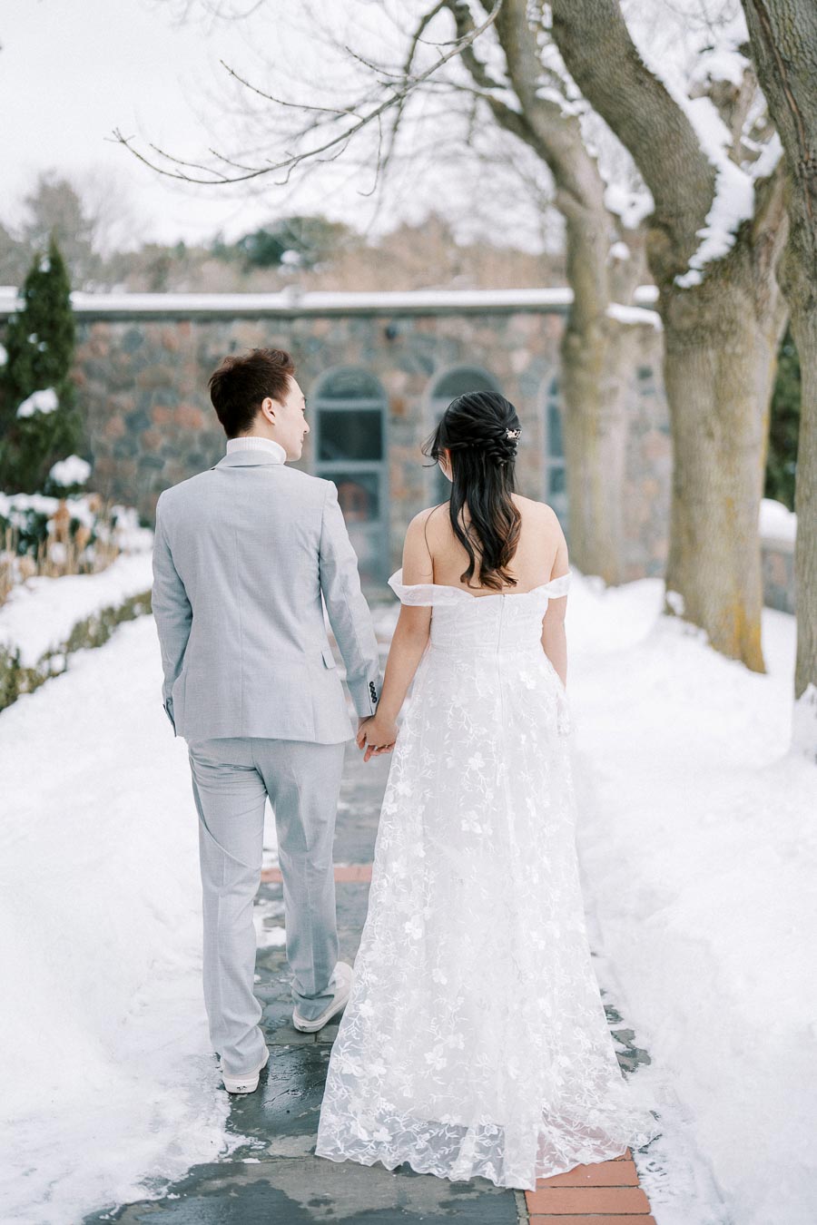 A couple holding hands, dressed in wedding attire, walking on a snowy path with trees and a stone building in the background.