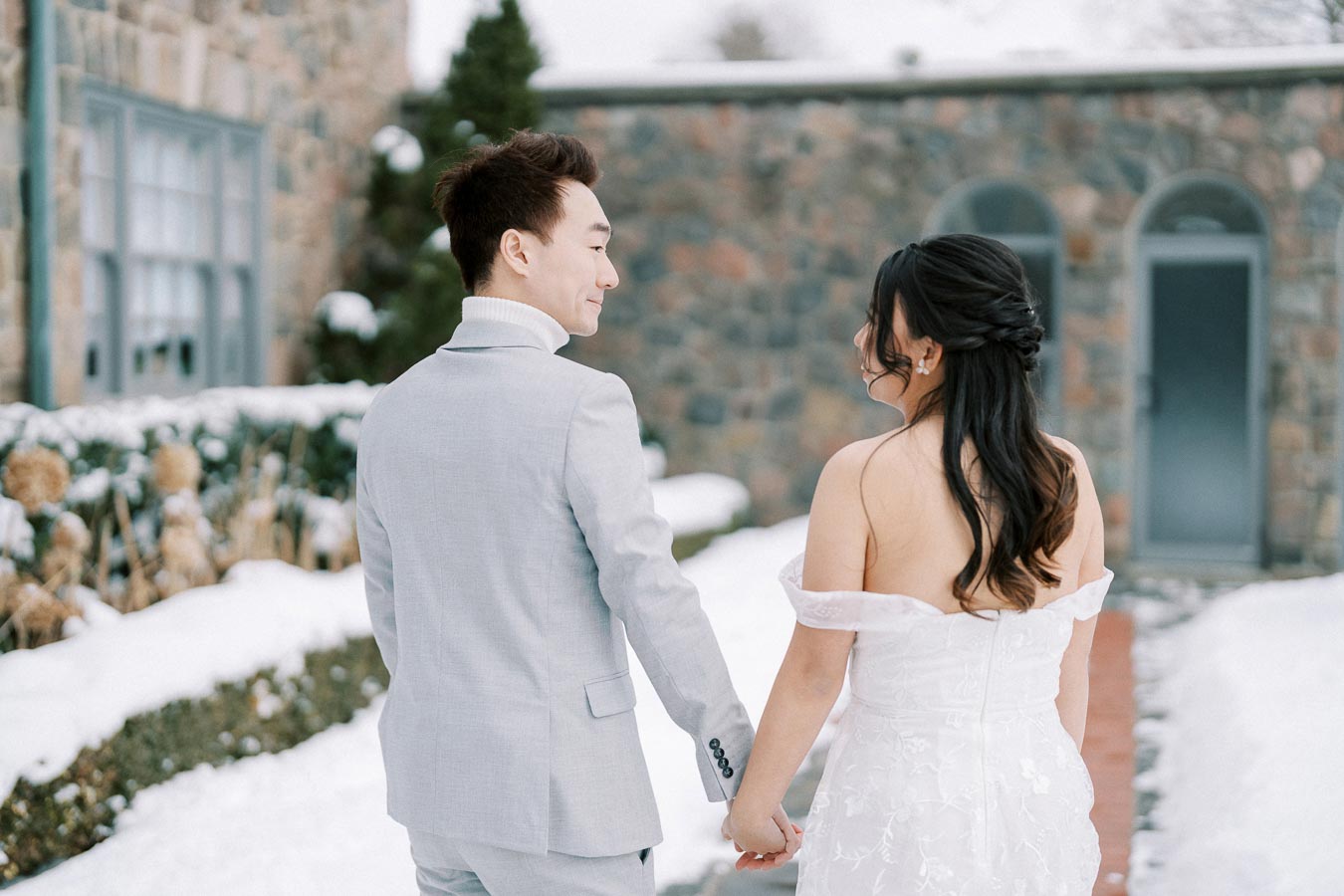 A couple holding hands in a snowy outdoor setting, both dressed in elegant wedding attire; the bride is wearing a white strapless gown and the groom is in a light grey suit, with a stone building in the background.