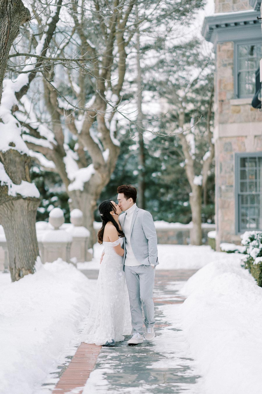 A couple in elegant winter attire shares a romantic kiss on a snow-covered path, surrounded by trees and a charming stone building in the background.