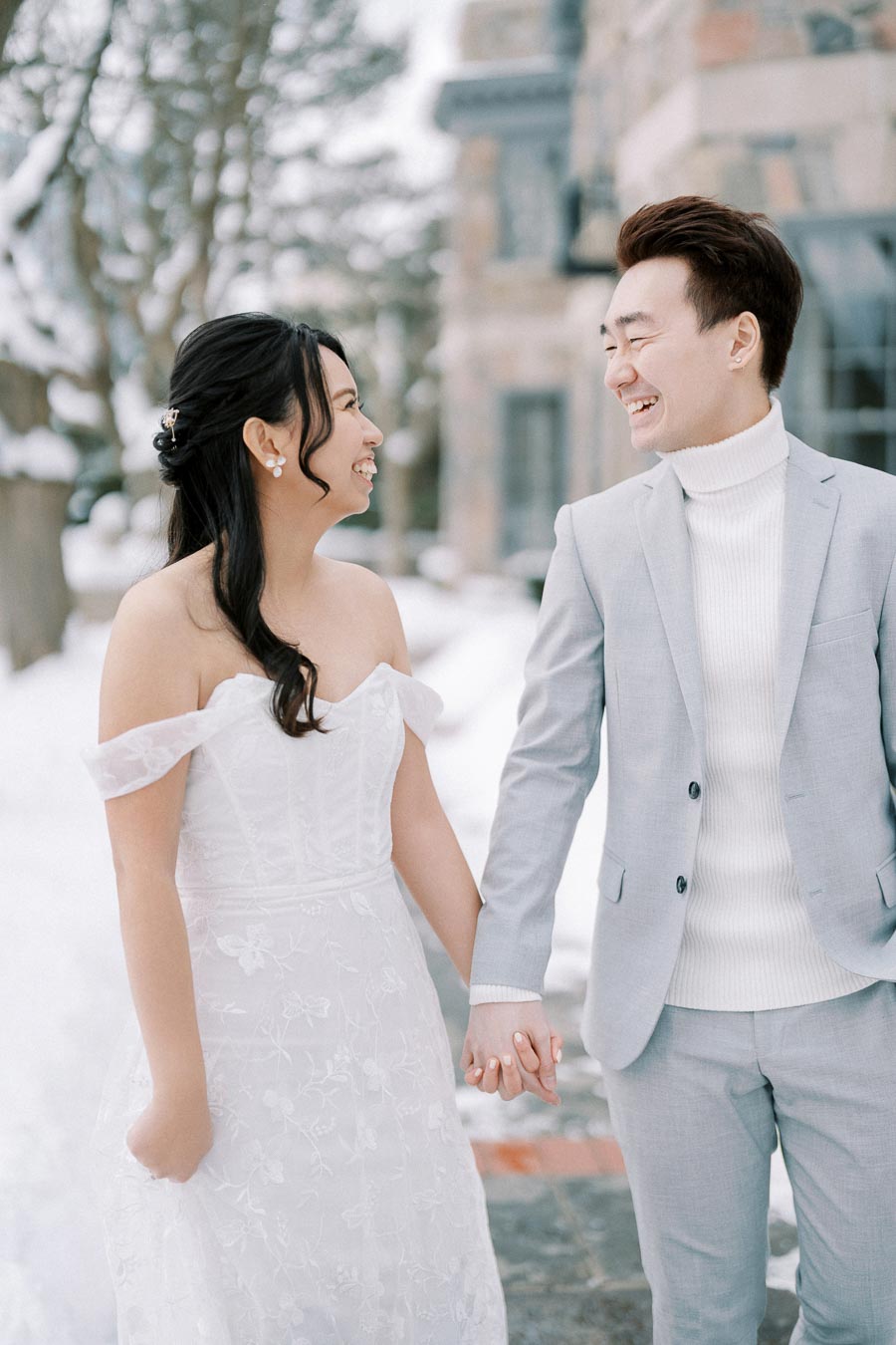 A couple smiling and holding hands, dressed in elegant wedding attire, with a snowy background and historical building in the distance.