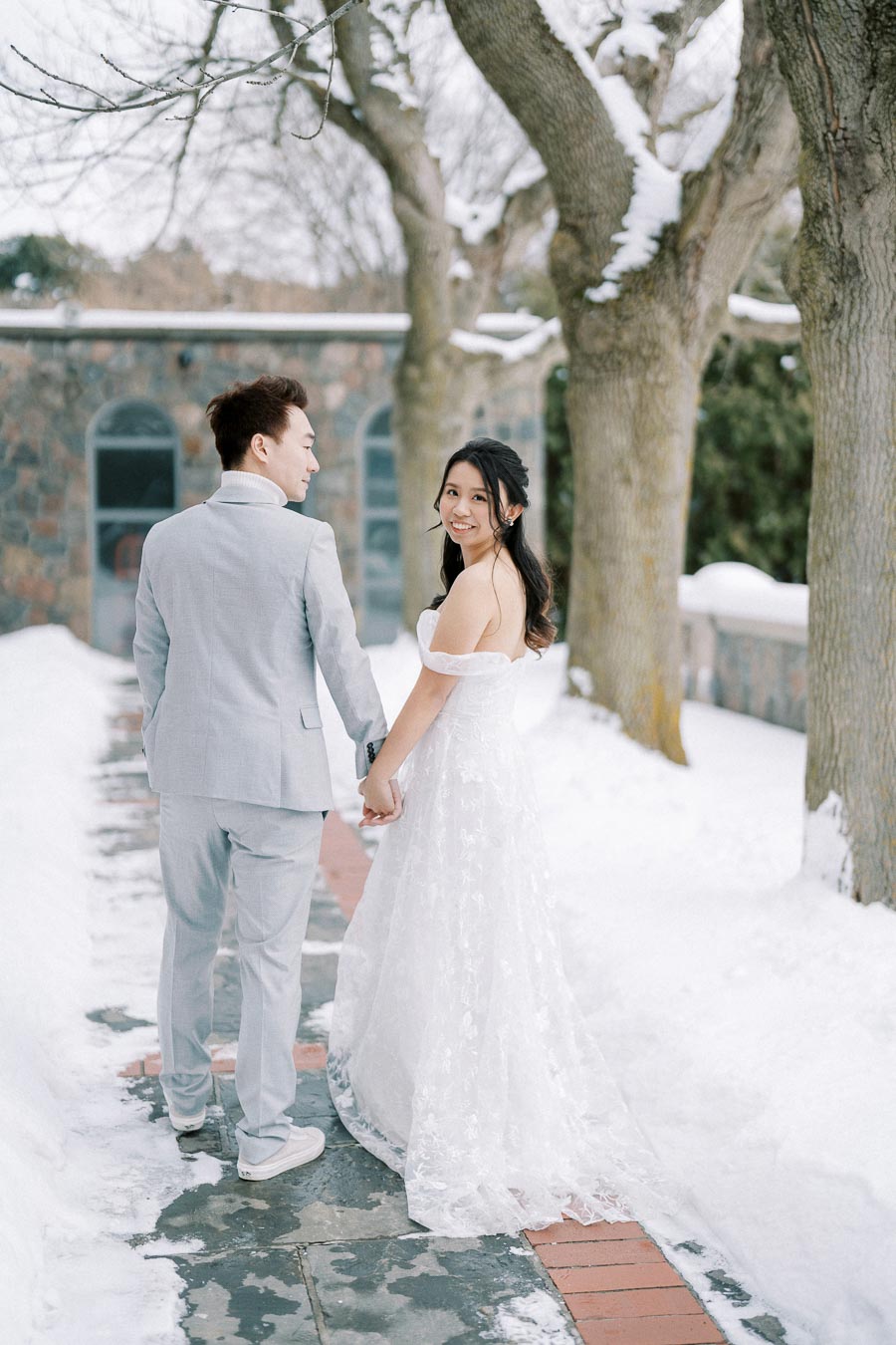A couple holding hands in winter wedding attire, walking along a snow-covered path lined with trees, with the bride in a white lace dress and the groom in a light gray suit.
