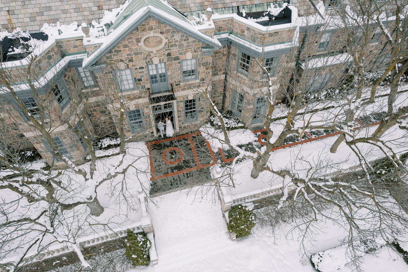 Aerial view of a large stone building surrounded by snow-covered trees and pathways, showcasing winter architecture and landscaping.