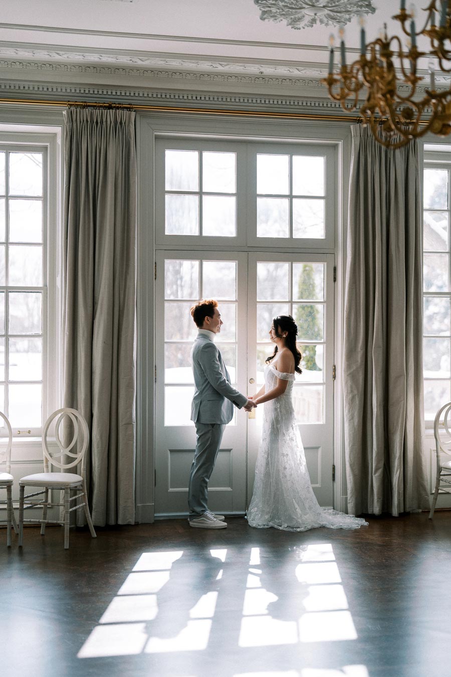 Elegant couple in wedding attire holding hands by a sunlit window in a luxurious, softly lit room with classic decor.