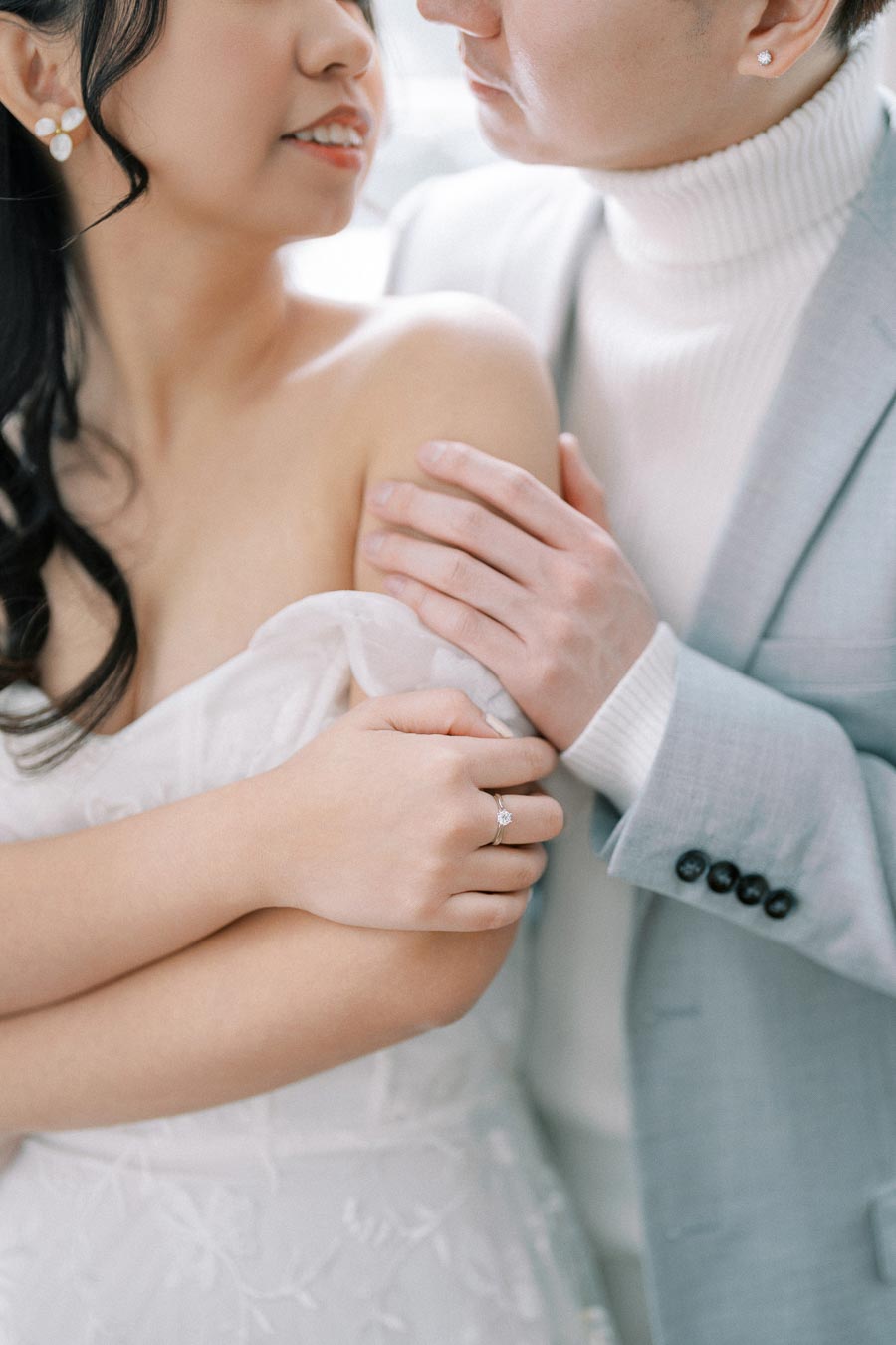 A romantic close-up of a couple embracing, with a focus on their hands and engagement ring. The groom is wearing a light blue suit and white turtleneck, while the bride is in an elegant off-shoulder wedding dress with floral lace details. This intimate moment captures love and connection.