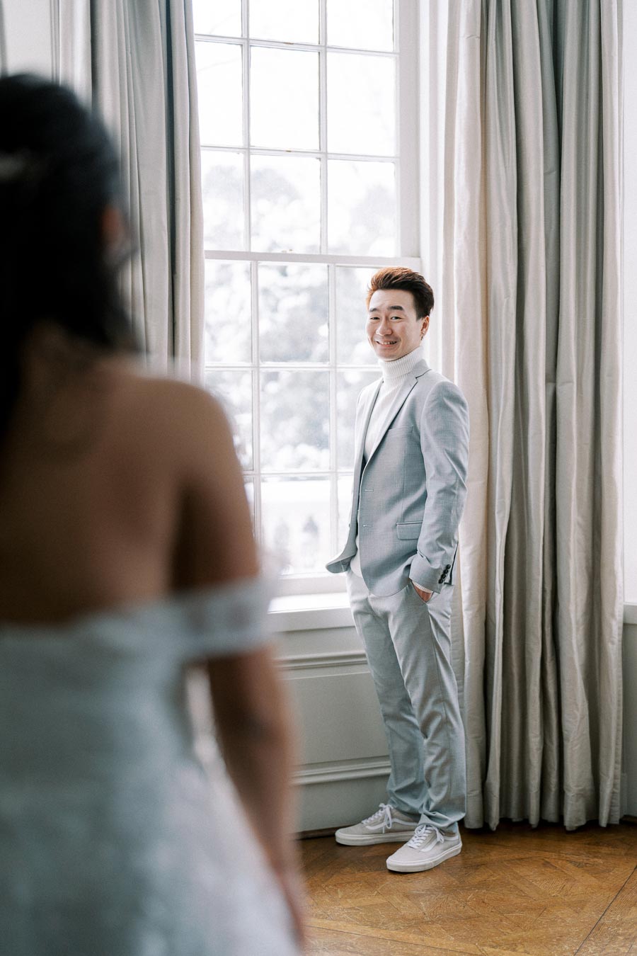 A groom in a light gray suit smiles while standing by a large window, with a blurred view of a bride in an off-shoulder wedding dress in the foreground, creating a romantic atmosphere in a softly lit room with elegant curtains.