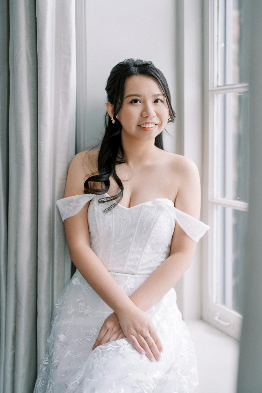 A bride in an elegant off-shoulder white wedding dress with floral lace details, sitting by a sunlit window with soft curtains, smiling gently at the camera.