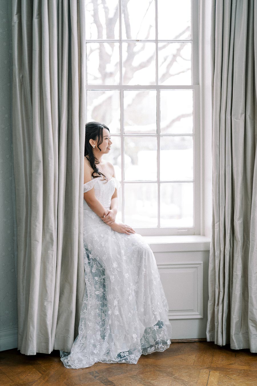 A bride in a lace off-the-shoulder wedding dress sits by a large window with soft light streaming in, creating an elegant and serene bridal portrait.