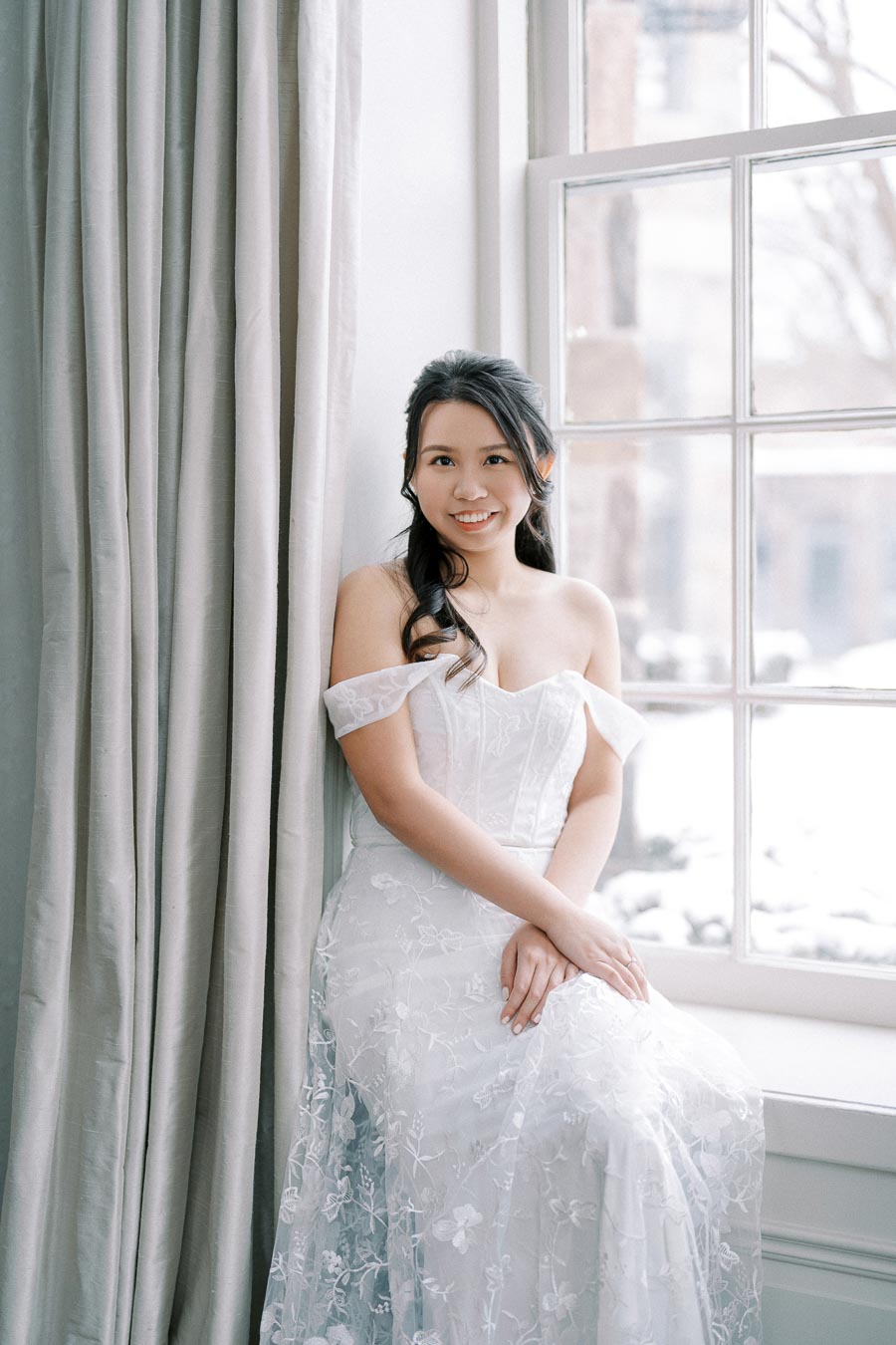 A bride in an elegant white off-shoulder wedding dress poses by a large window, with a serene expression and softly curled hair, creating a romantic and classic bridal portrait setting.