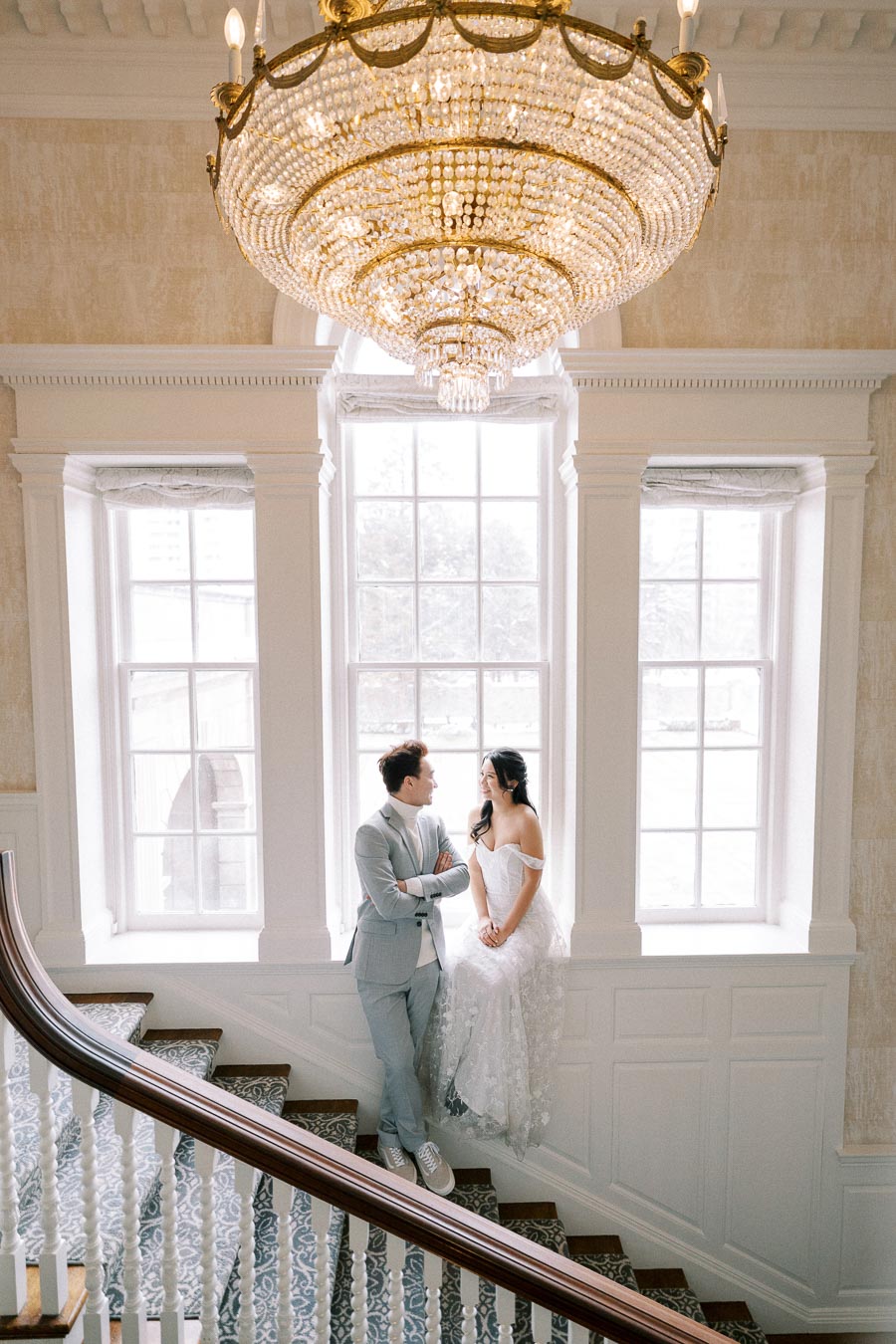 Wedding couple posing on a staircase under an elegant crystal chandelier, surrounded by large windows with natural light streaming in.