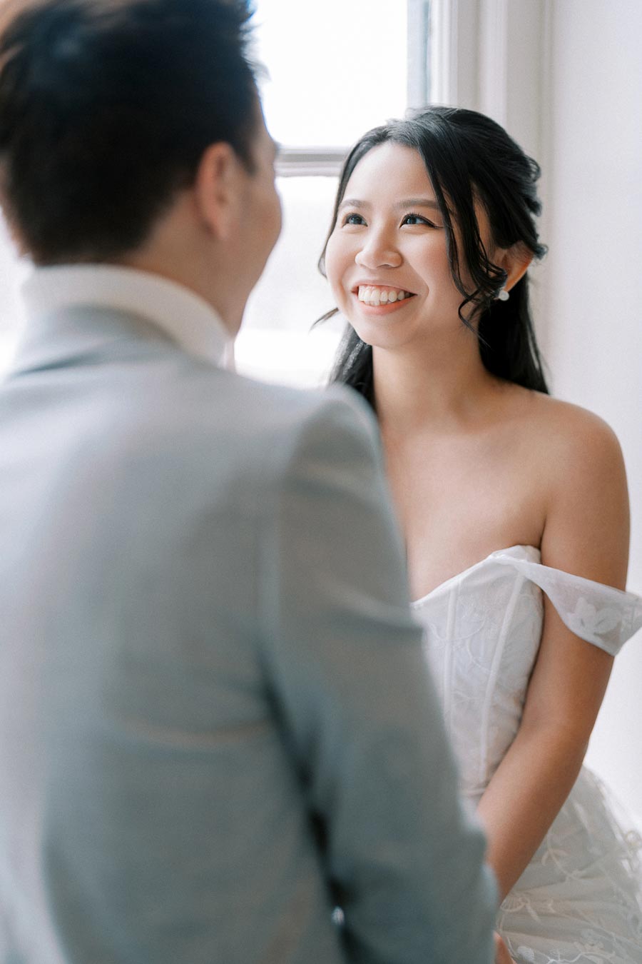 A joyful bride in a white off-shoulder wedding dress smiles at her partner inside a softly lit room.