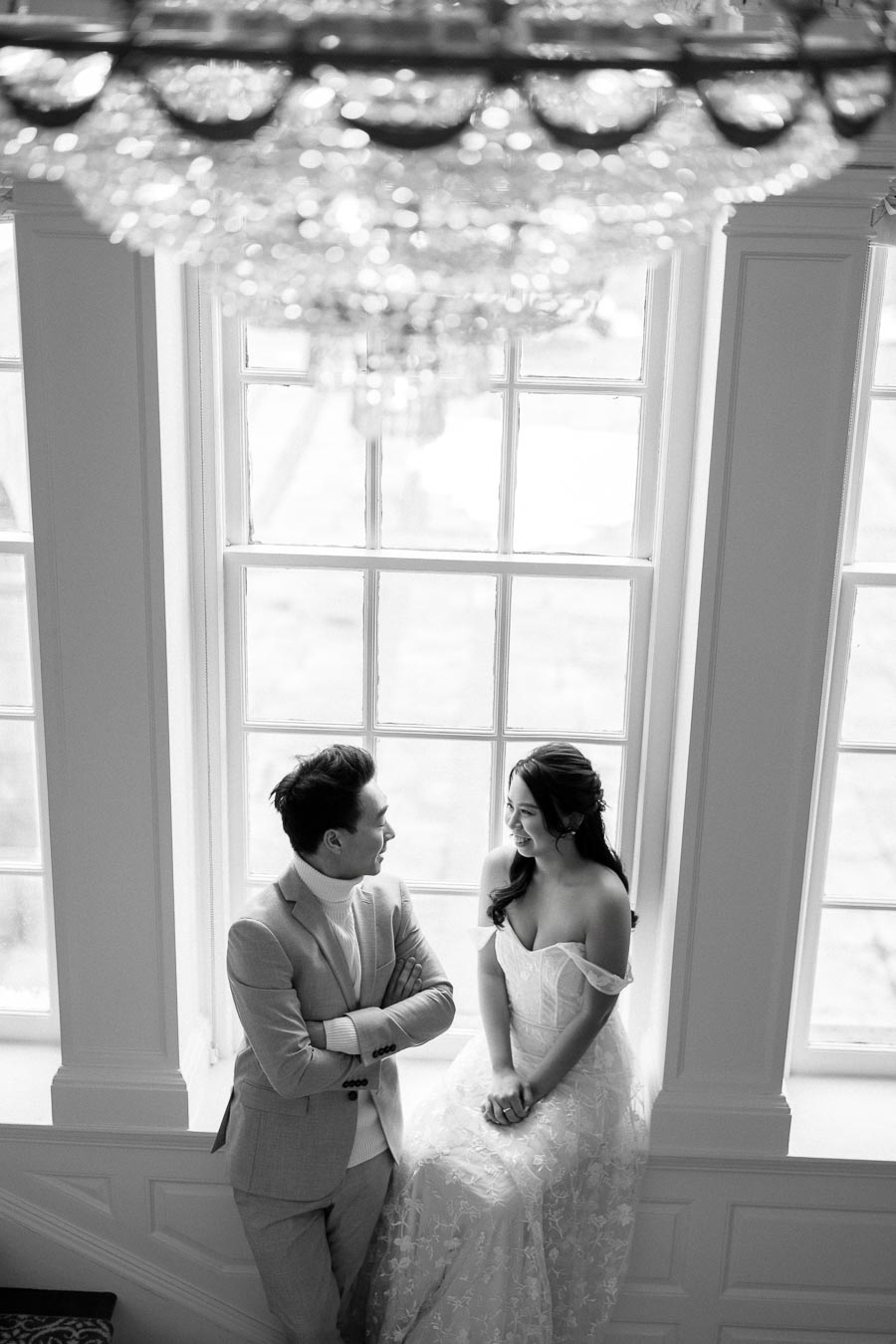 Black and white photo of a couple in wedding attire smiling at each other while sitting on a window sill under a chandelier in a beautifully lit room.