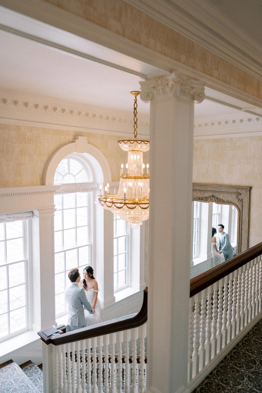 Elegant wedding couple standing on a grand staircase next to vintage windows under a luxurious chandelier, embracing in a romantic setting.